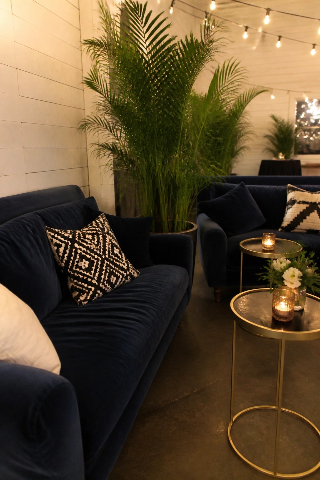 Navy blue velvet sofas with black and white pillows sit beside gold tables holding candles and white flowers, surrounded by tall palm plants under string lights in a white walled venue.