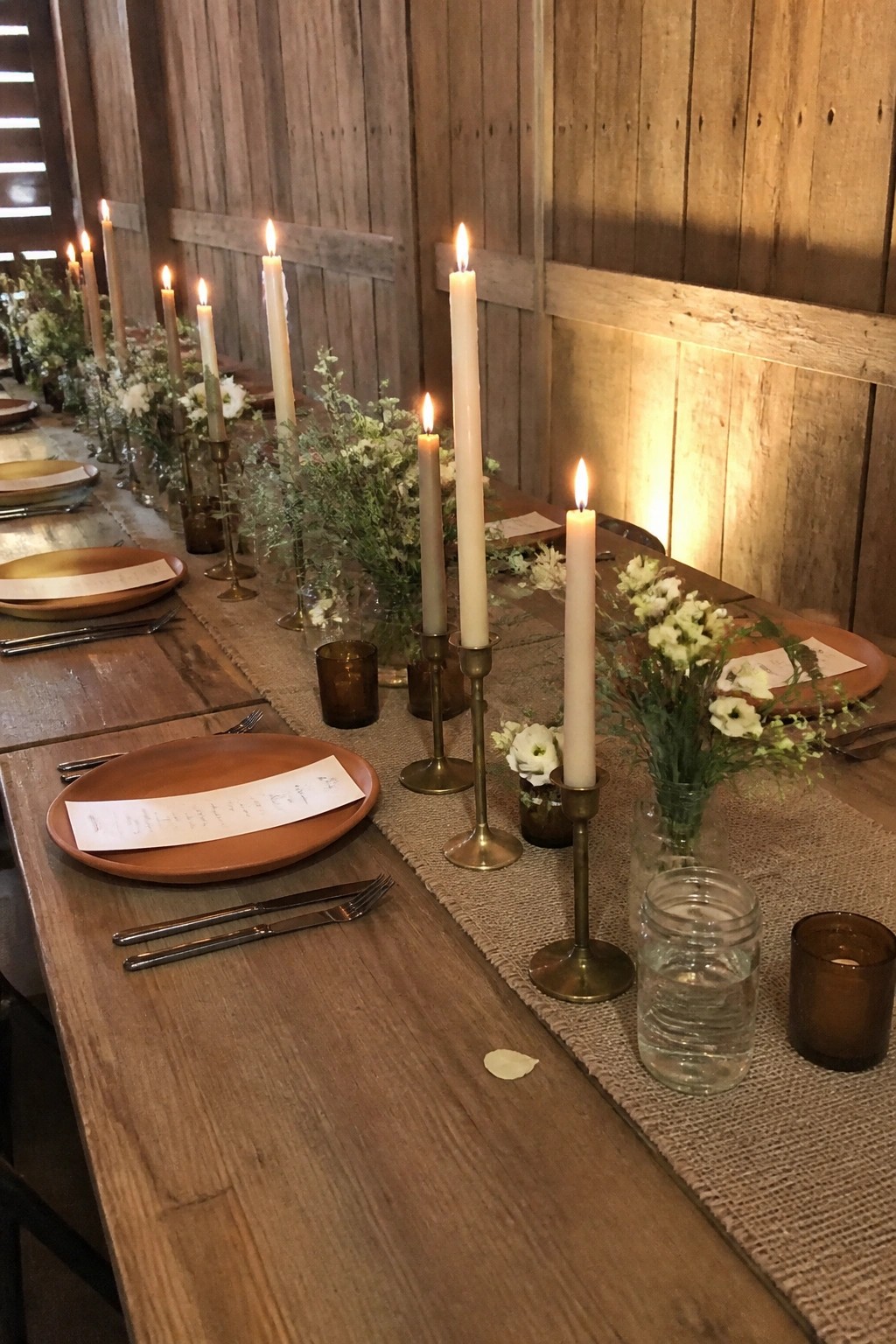 Long rustic wedding table with burlap runner, terracotta plates, brass candlesticks holding lit white candles, small floral arrangements in glass jars, and wooden barn walls in warm candlelight.