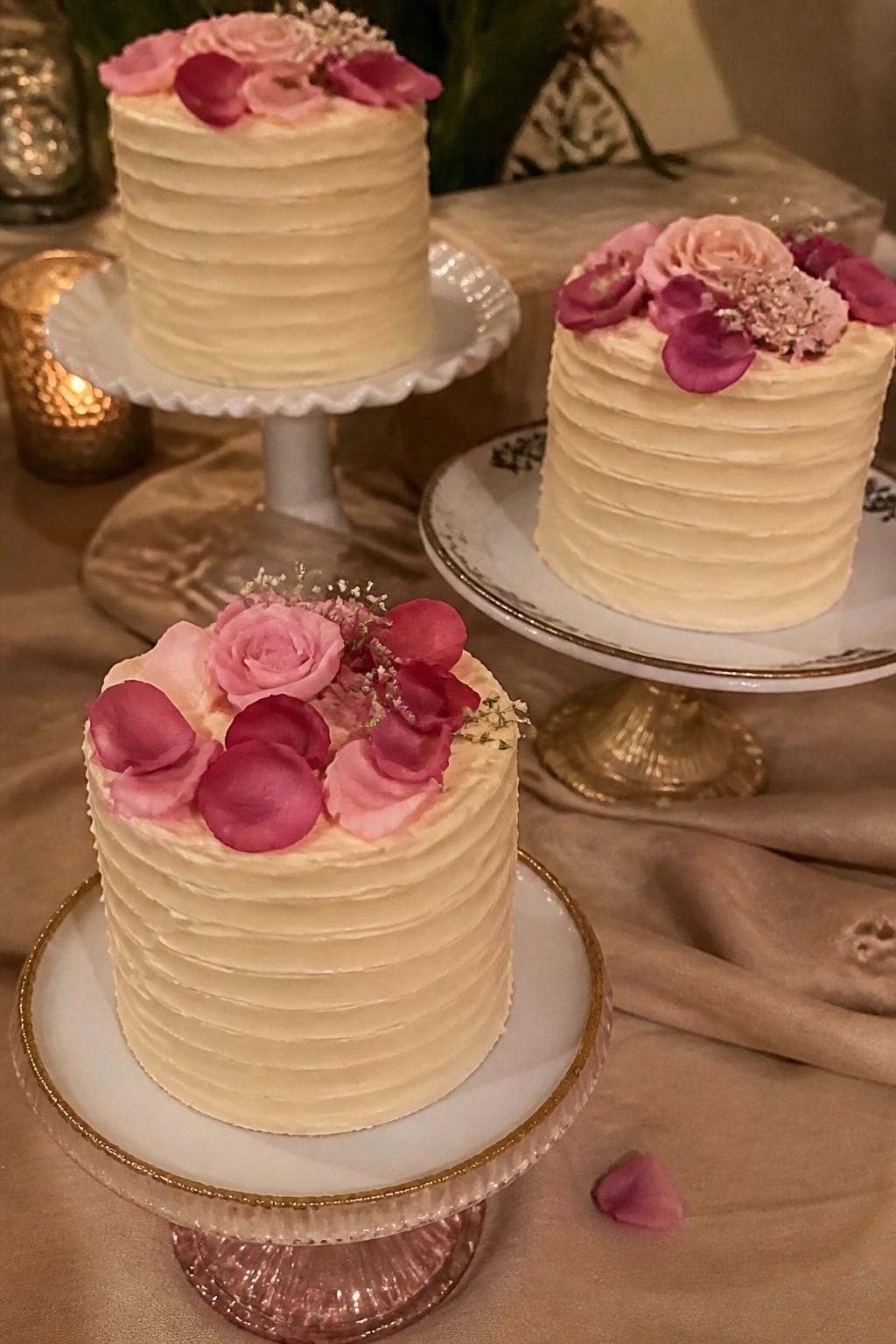 Three cream frosted cakes topped with pink rose petals and small flowers displayed on decorative stands in a softly lit wedding setting