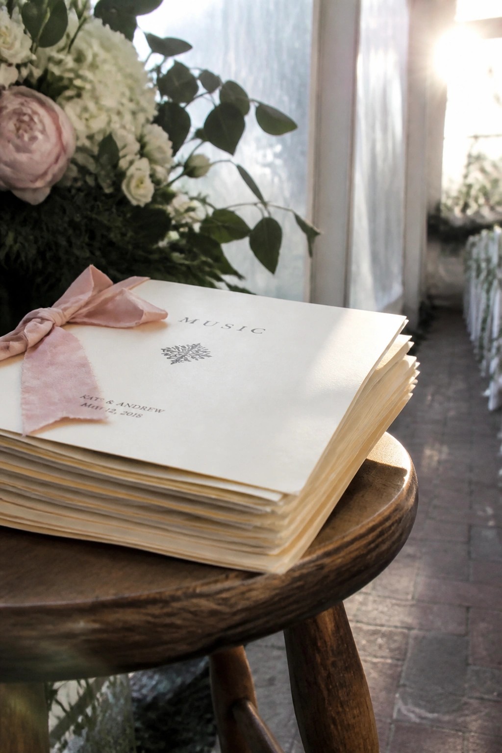 Stack of cream wedding music programs tied with pink velvet bow on wooden stool next to floral arrangement in bright sunlit venue