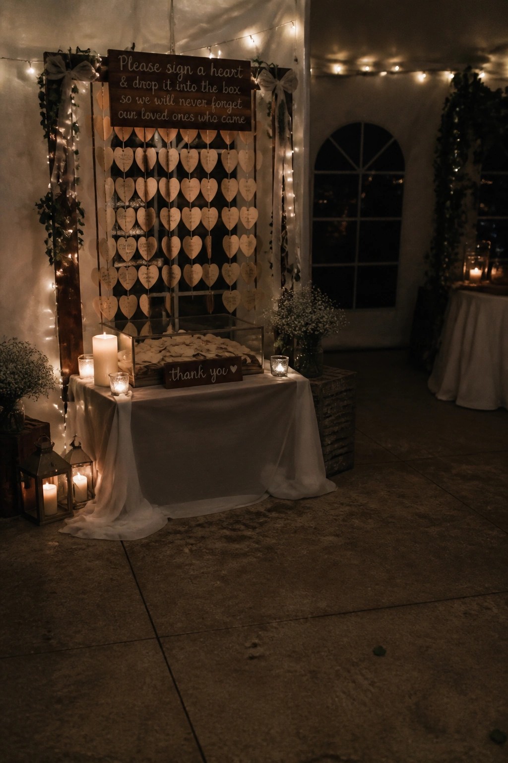 Nighttime wedding memorial display with rows of signed wooden hearts on a lit frame and a glass box on a candlelit table for collecting them.