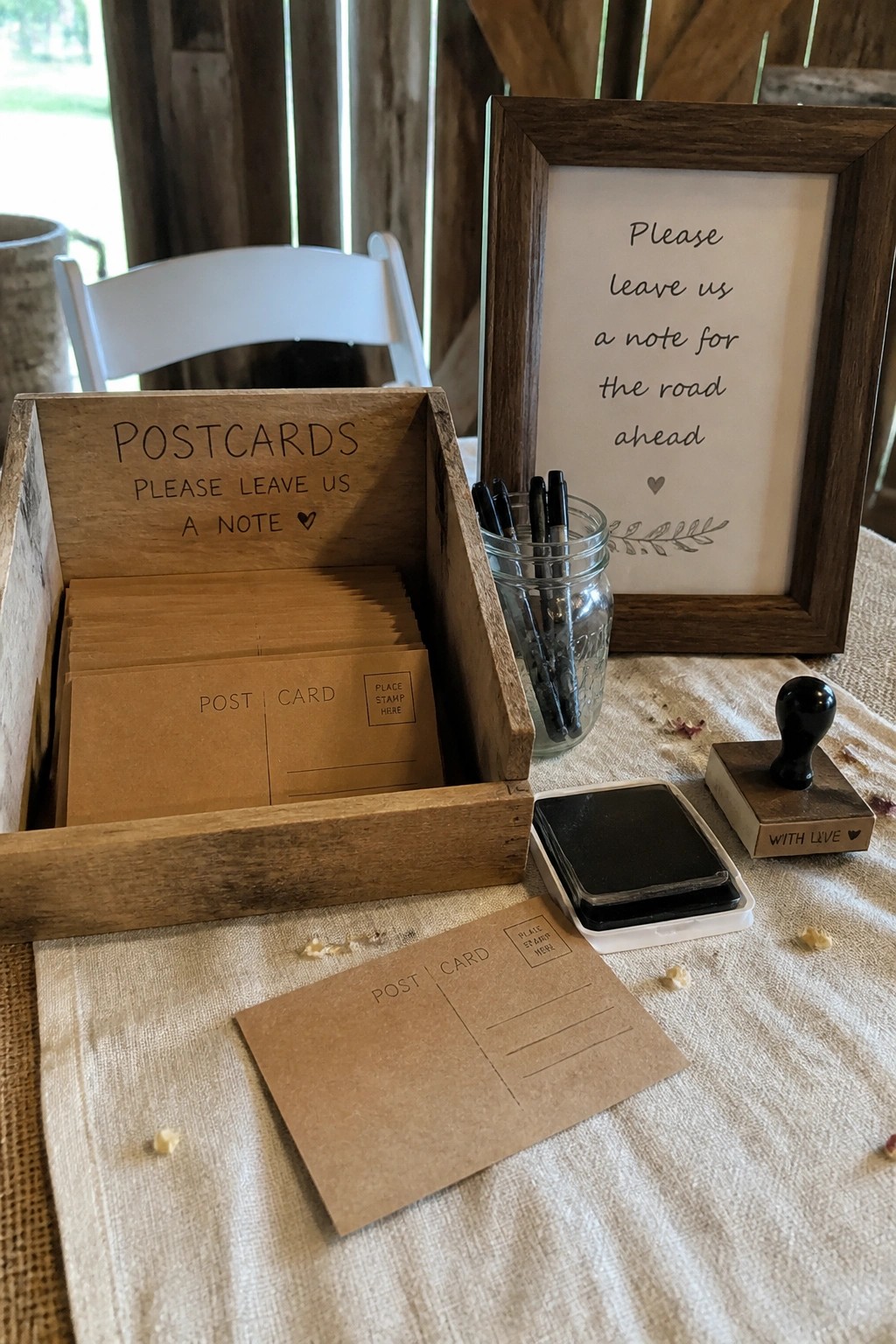 Rustic wooden box of blank postcards with pens, ink stamp, and framed sign inviting wedding guests to leave notes on a linen table.