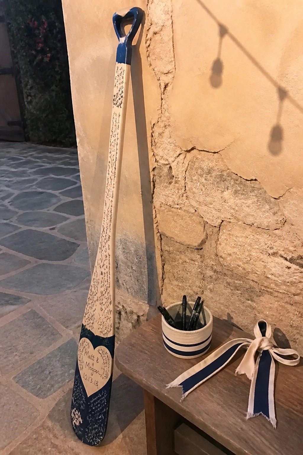 A decorated wooden paddle signed by wedding guests leans against a stone wall with markers and a ribbon on a nearby table.