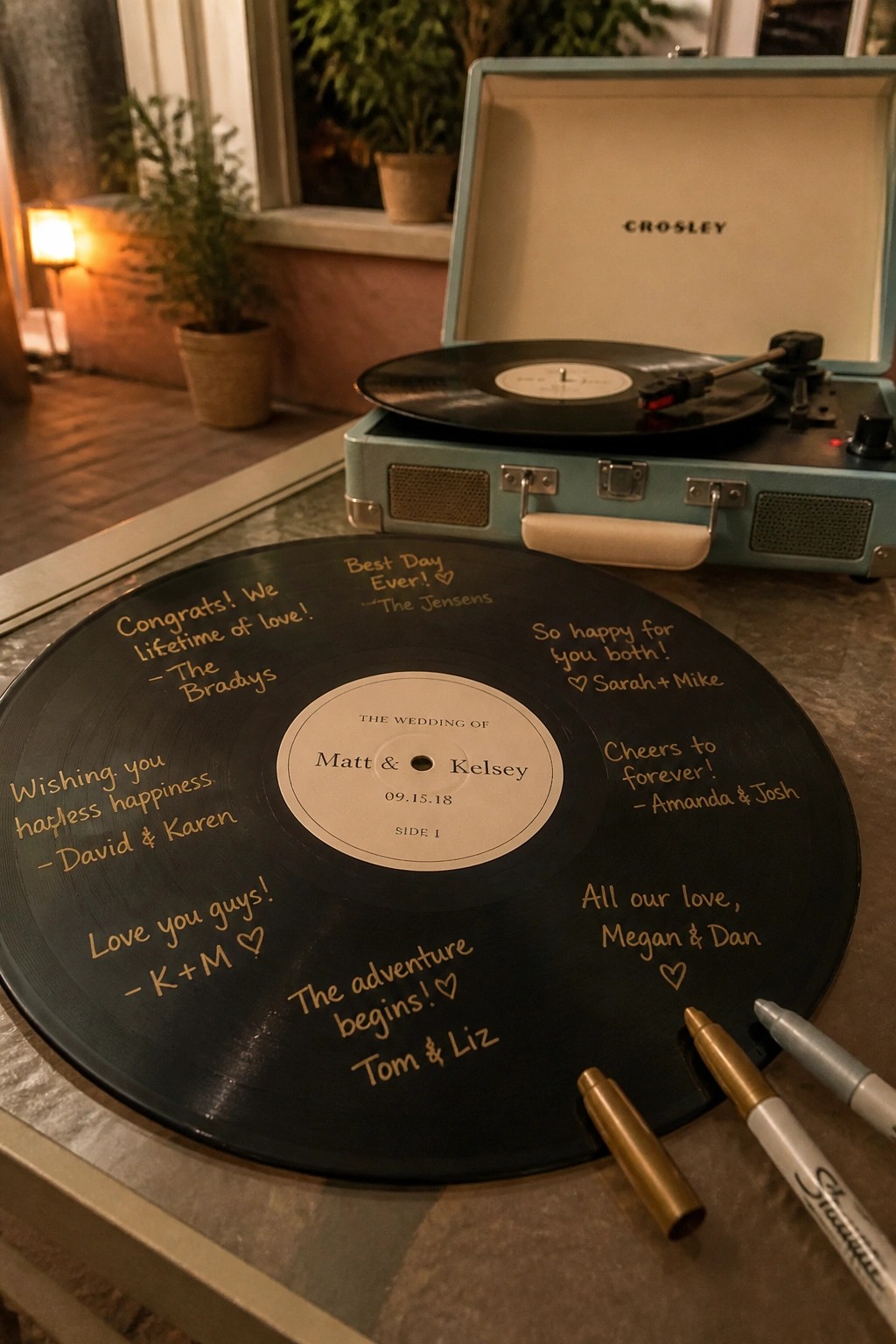 A black vinyl record signed with gold and silver messages from wedding guests sits in front of a turquoise Crosley record player with markers nearby.