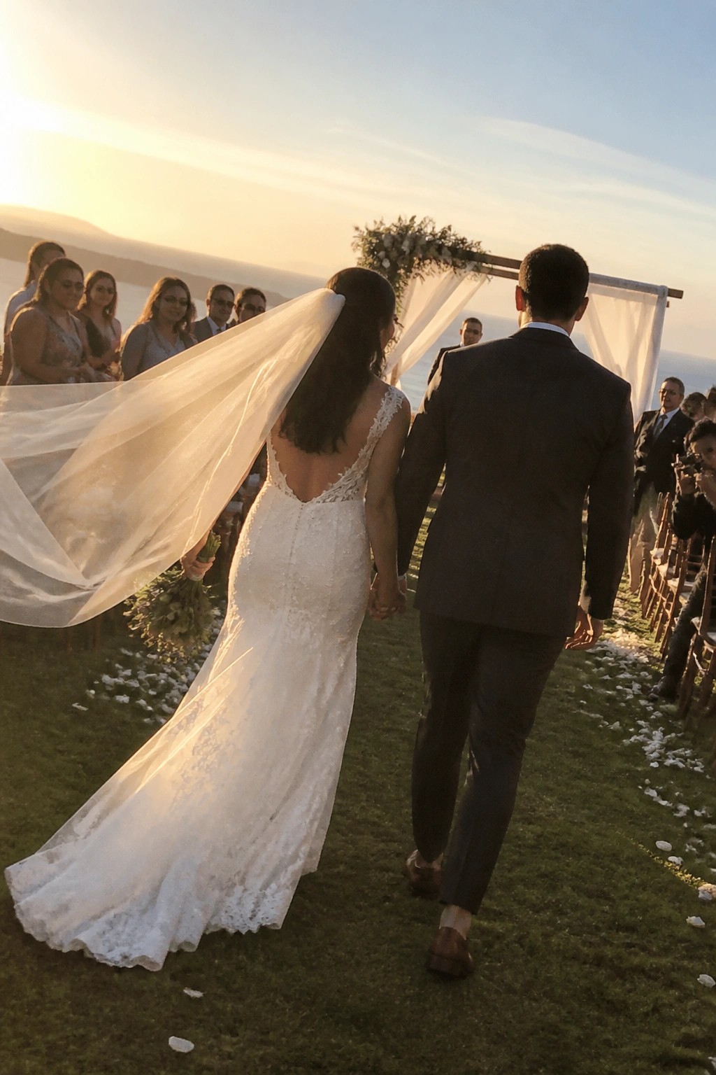 Bride in lace gown with flowing veil walks hand in hand with groom toward a floral arch overlooking the ocean at sunset