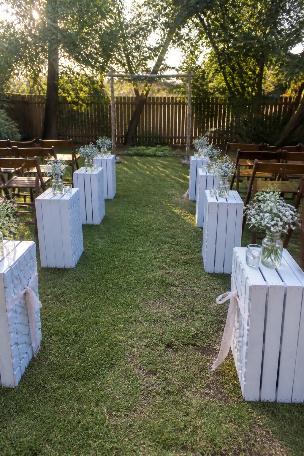 Outdoor wedding aisle lined with white wooden crates holding baby's breath flowers and candles on green grass with wooden chairs and an arch at sunset