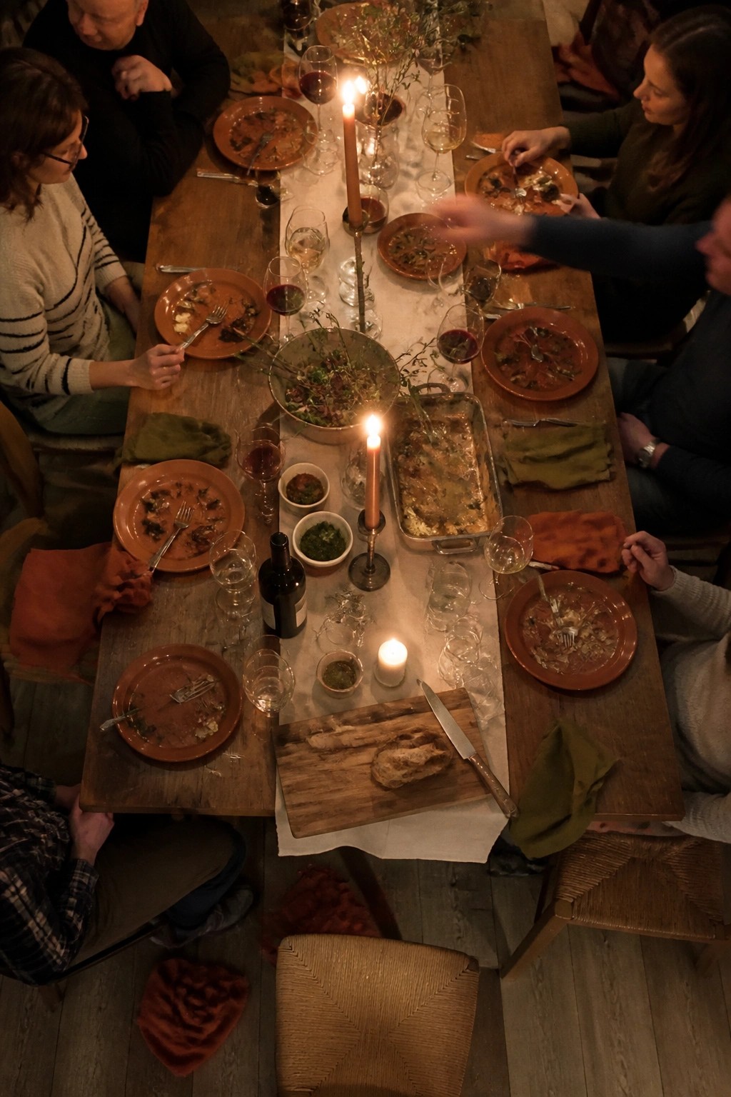 Top down view of a rustic boho wedding dinner table with terracotta plates, lit candles, olive napkins, wine glasses, and guests seated around a long wooden table in warm evening light.