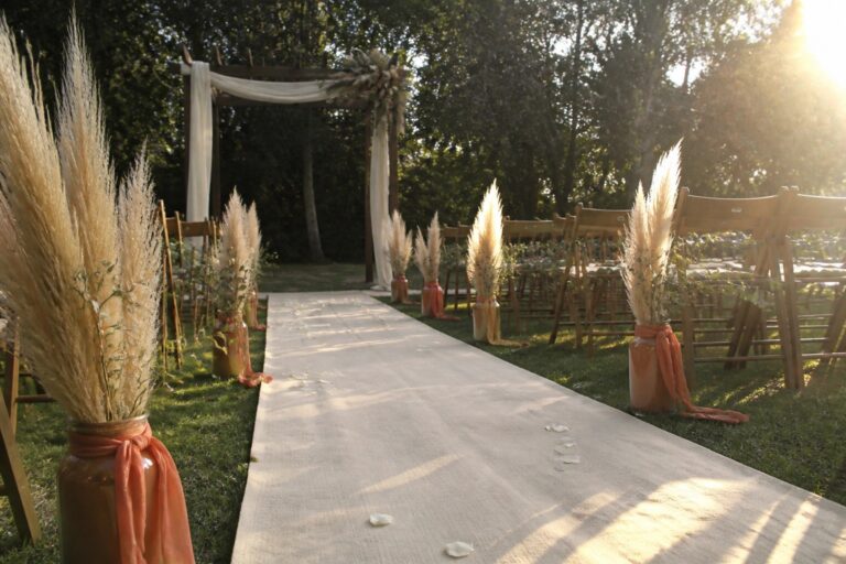 Outdoor boho wedding aisle lined with terracotta jars of pampas grass, cream runner with petals, wooden chairs with greenery, and a fabric draped arch at sunset.