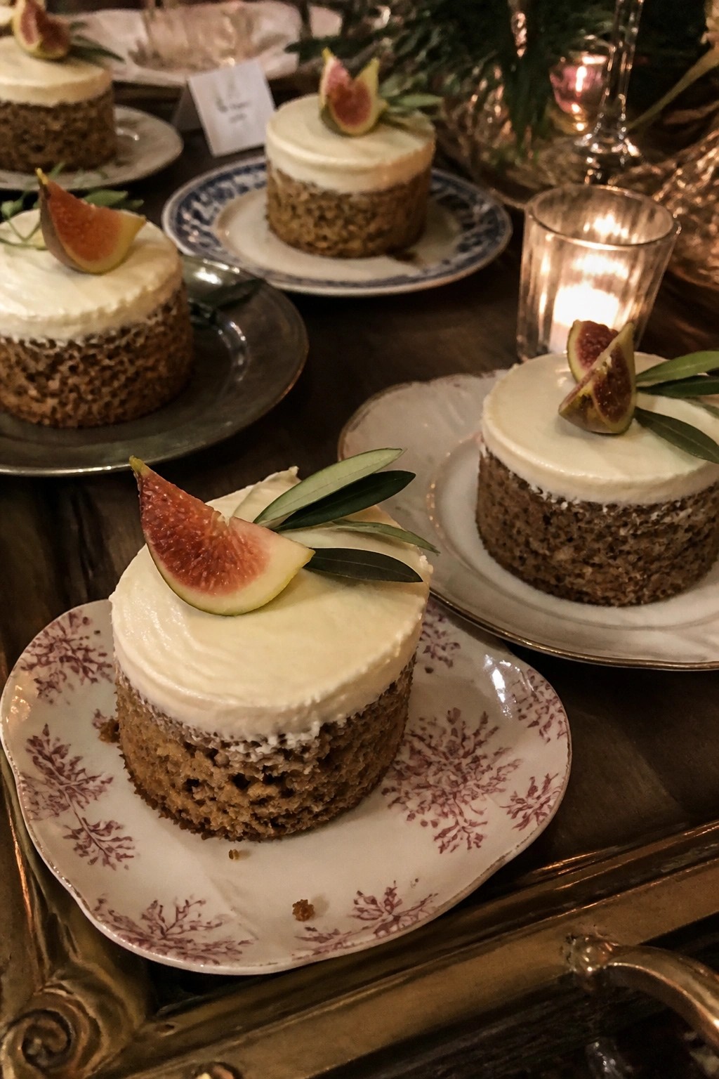 Small round cakes with white frosting topped with fig slices and olive leaves on vintage floral plates atop a wooden table with glowing candles in a boho wedding setting.
