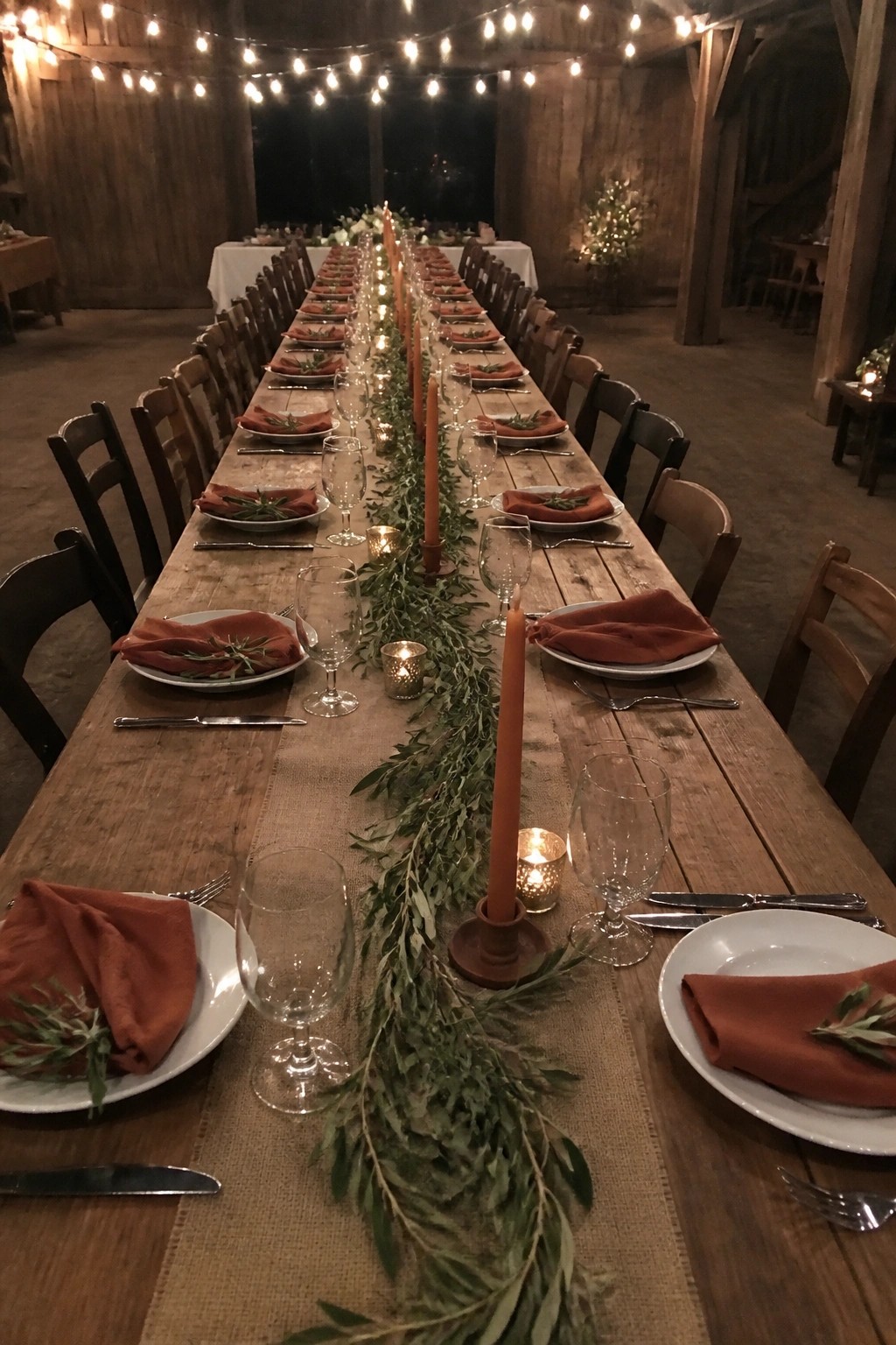 Long wooden wedding table in a barn with terracotta napkins, olive greenery runner, tall candles, and string lights overhead.