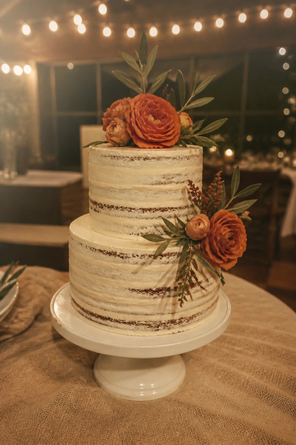 Two tier naked wedding cake with white frosting terracotta flowers and olive greenery on a white stand over burlap in a string lit venue