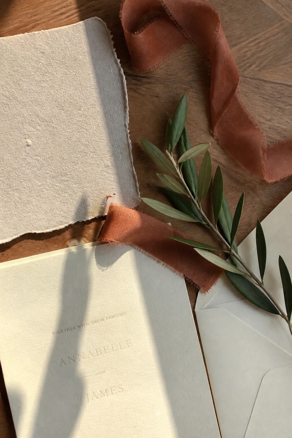 Cream wedding invitation with terracotta ribbon and olive branch on wooden table in soft light