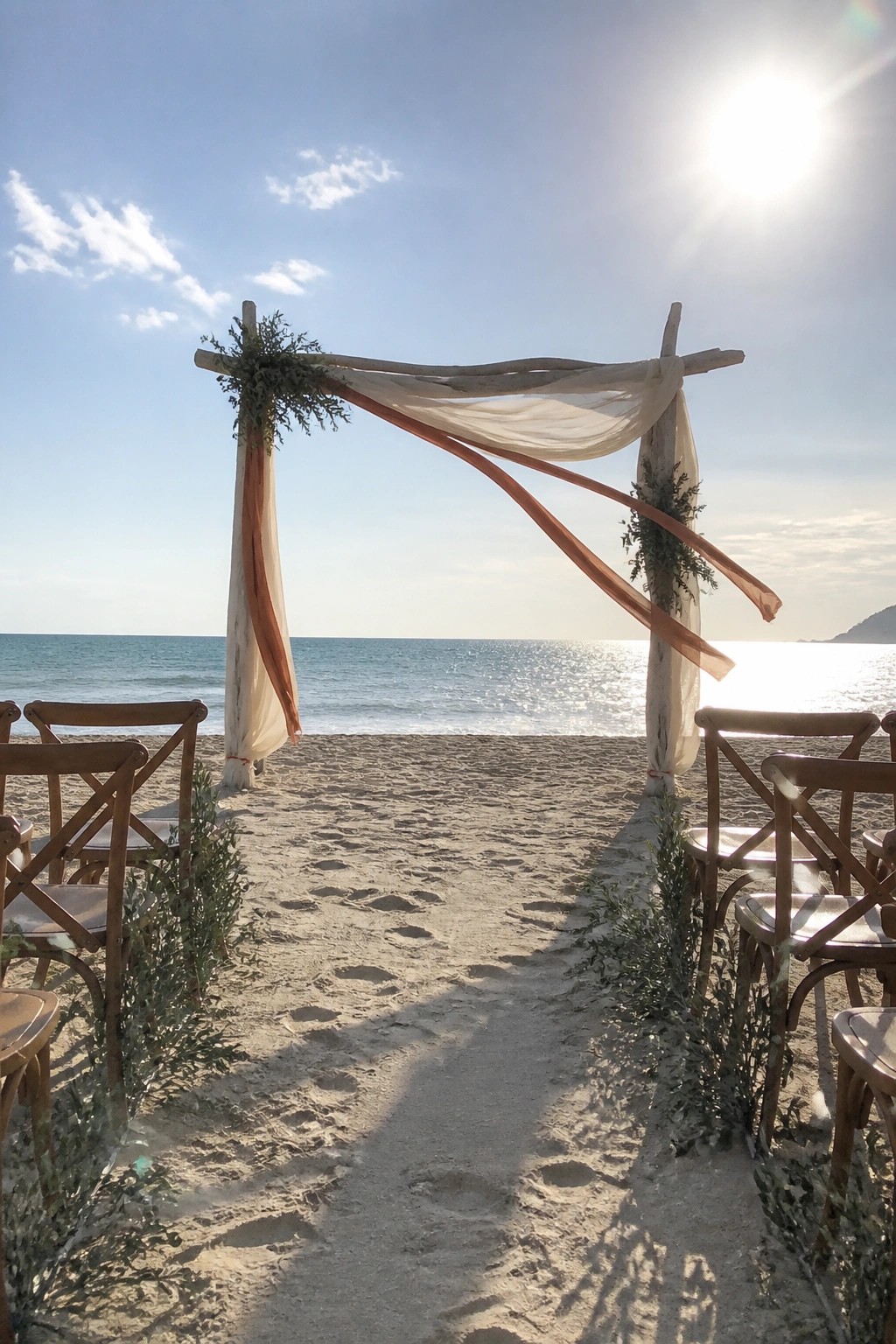 Rustic wooden wedding arch draped in cream and terracotta fabrics with olive greenery on a sandy beach facing the ocean with wooden chairs along the aisle under bright sunlight.