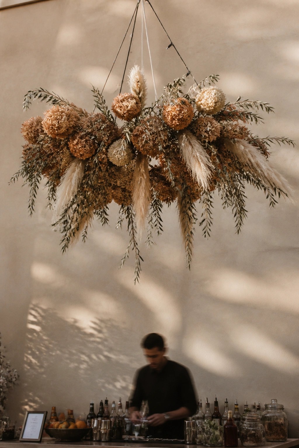Hanging boho wedding floral arrangement with pampas grass terracotta hydrangeas and olive greenery suspended above a bar with a bartender in the background