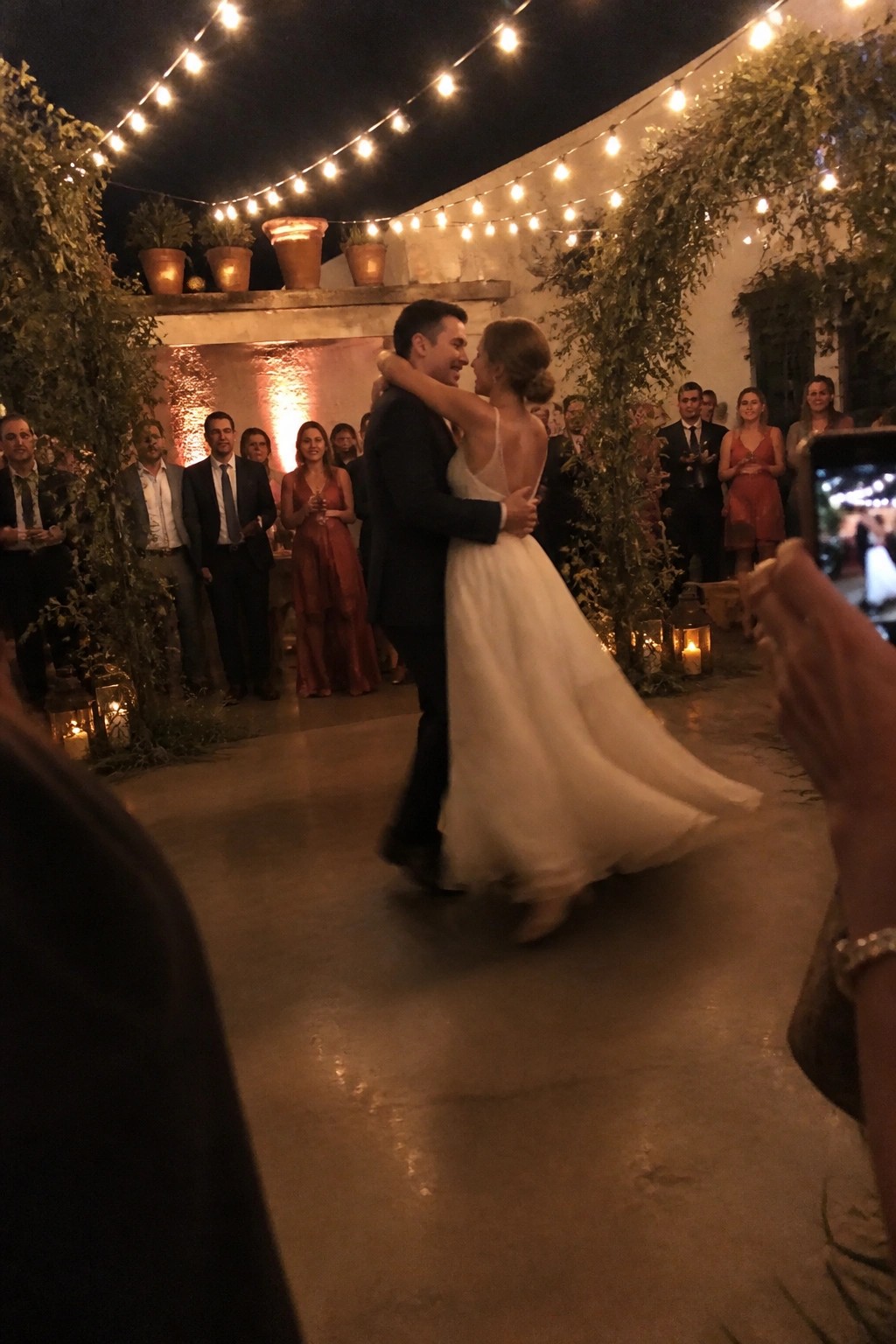 Bride in cream dress and groom in dark suit dance at night in a courtyard with string lights overhead and guests in terracotta dresses watching.