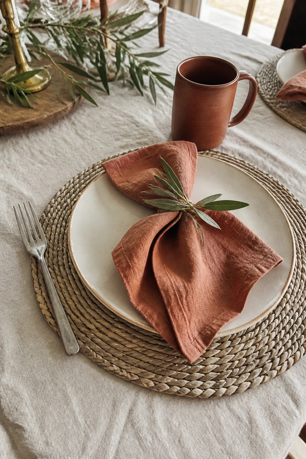Close up of a modern boho wedding place setting with a folded terracotta napkin and olive sprig on a cream plate atop a woven charger next to a silver fork and terracotta mug on a sand linen tablecloth.