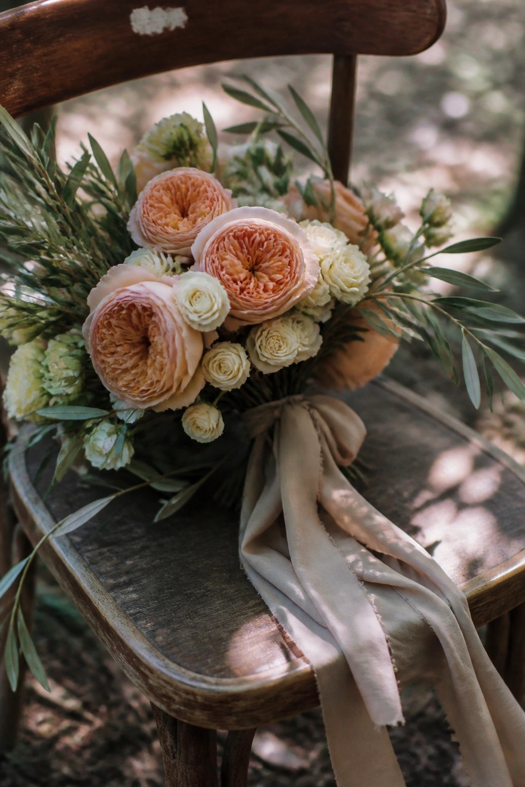 A lush wedding bouquet of peach terracotta roses and cream flowers with olive greenery tied by a sand ribbon sits on a rustic wooden chair outdoors.