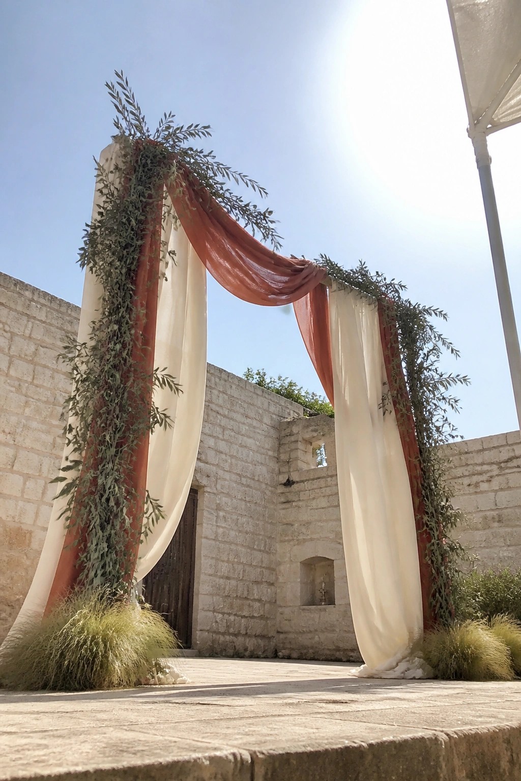 A boho wedding arch draped in cream and terracotta fabric with olive greenery stands against stone walls under a sunny blue sky