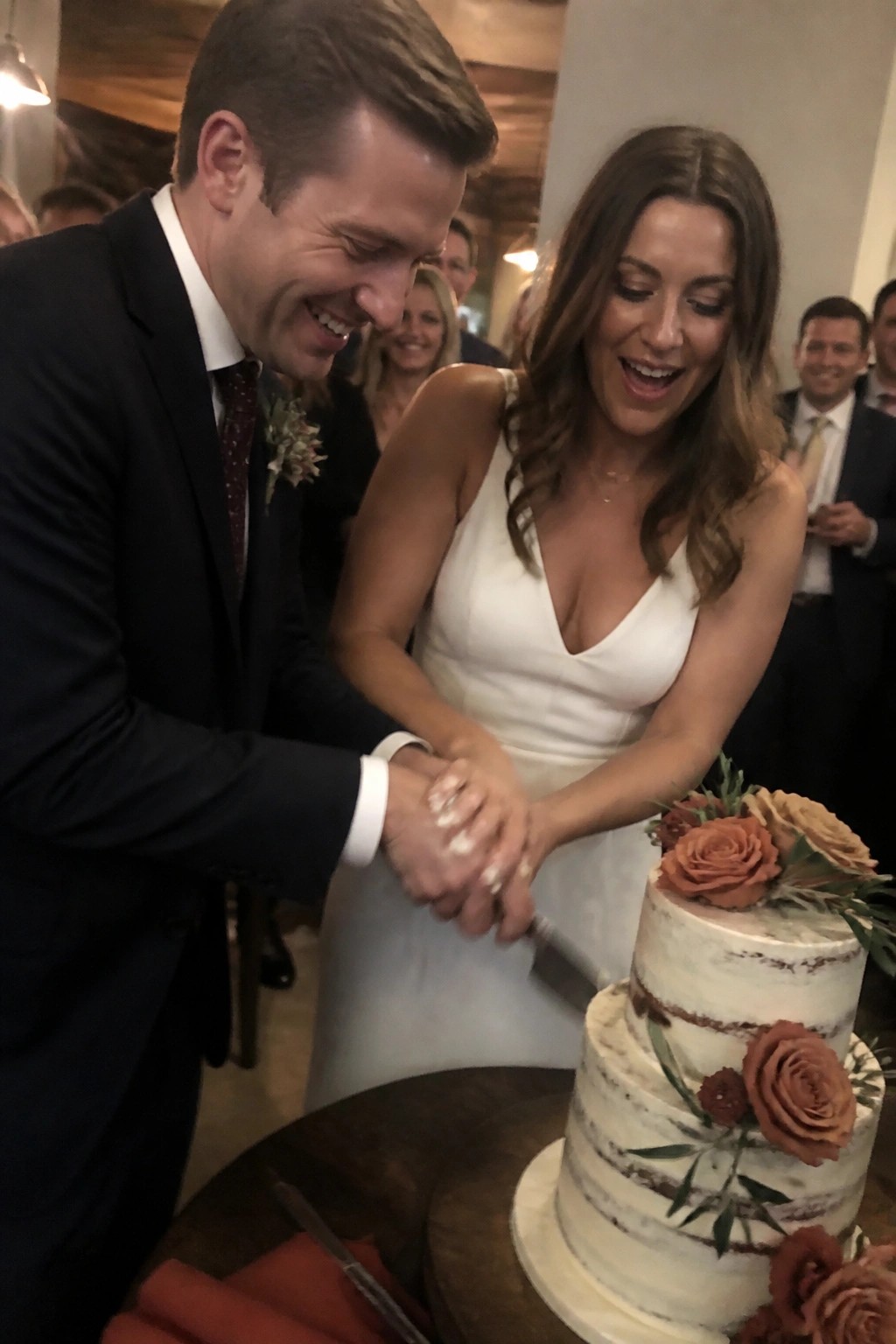 Bride and groom laughing while cutting a cream wedding cake decorated with terracotta roses and olive leaves at a rustic venue