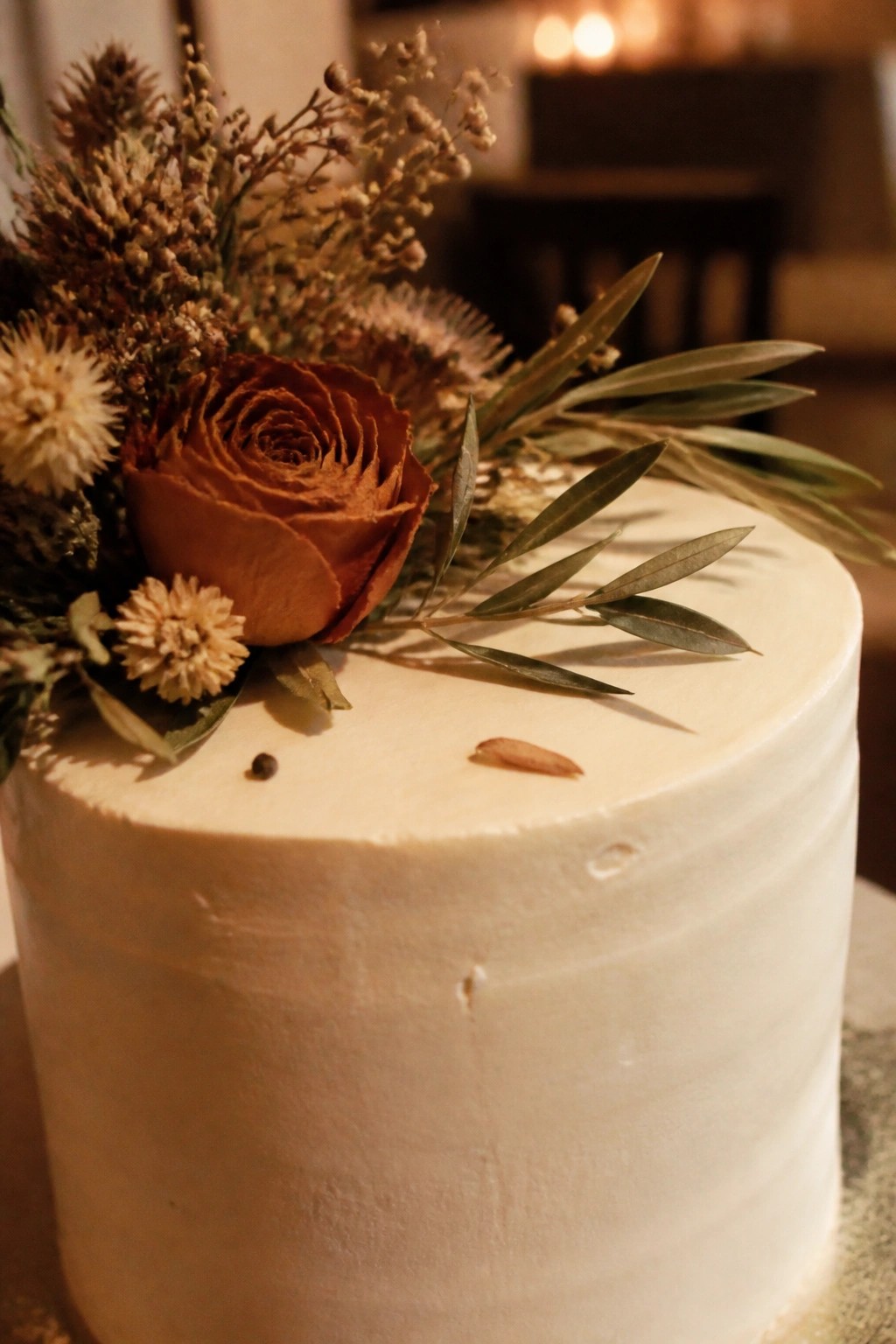 Close up of a single tier cream wedding cake topped with a large dried rust rose olive leaves and assorted dried flowers in beige tones under warm lighting.