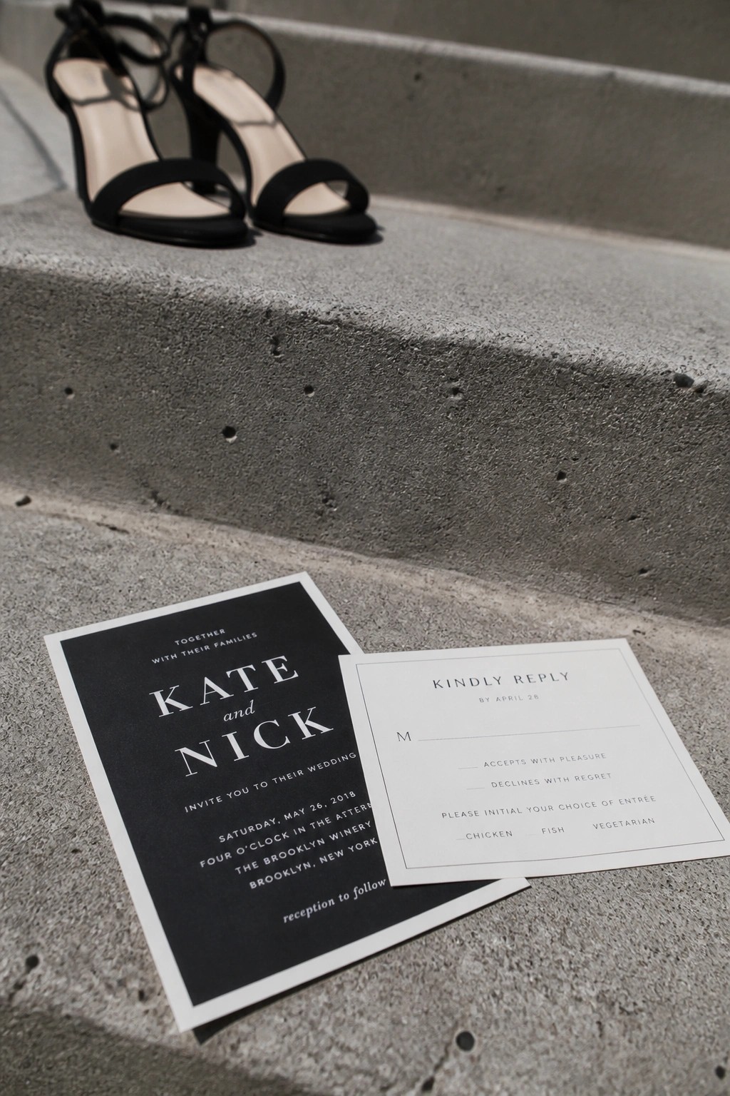 Black and white wedding invitation for Kate and Nick placed on concrete steps beside black high heeled shoes with a reply card on top