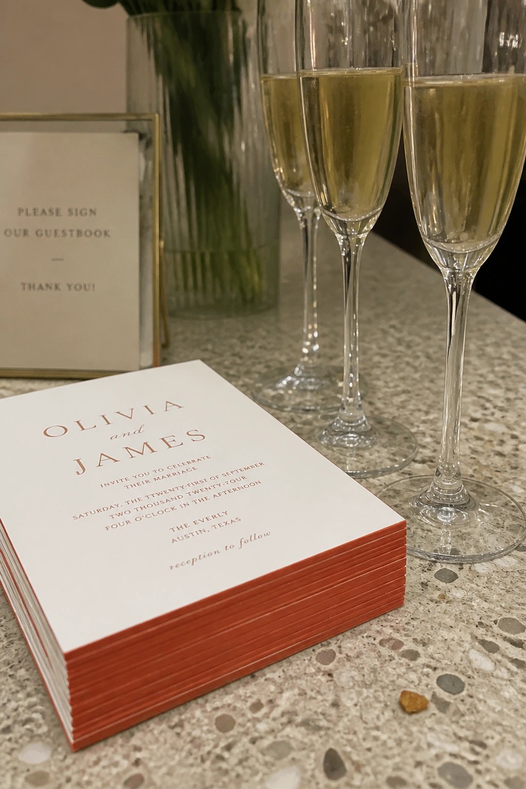 Stack of white wedding invitations with bright orange edges and gold text for Olivia and James on marble counter beside champagne flutes and guestbook sign