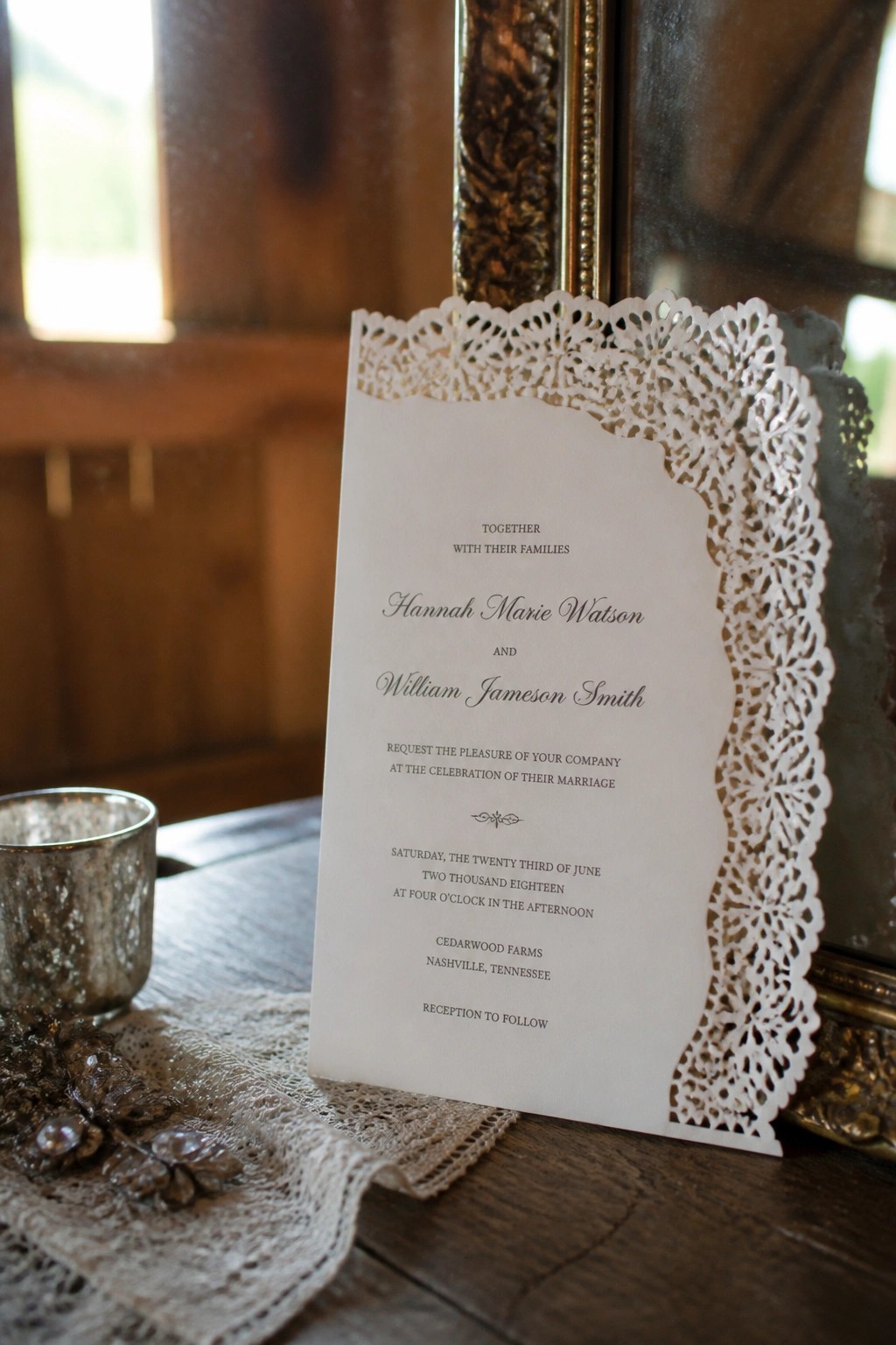 Close up of a white wedding invitation with ornate lace paper border displayed on dark wood with lace fabric and a small metallic glass.