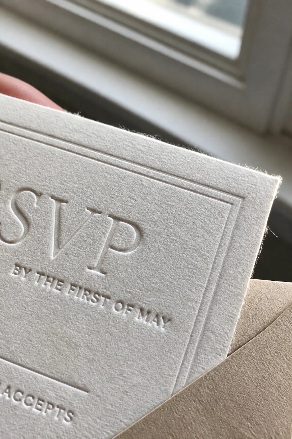Close up of a white embossed RSVP card with elegant typography held near a window in soft natural light.