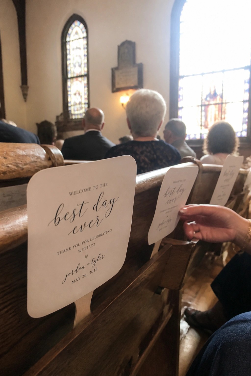 White wedding pew cards with black script reading best day ever rest on dark wooden church pews in a sunlit sanctuary with stained glass windows.