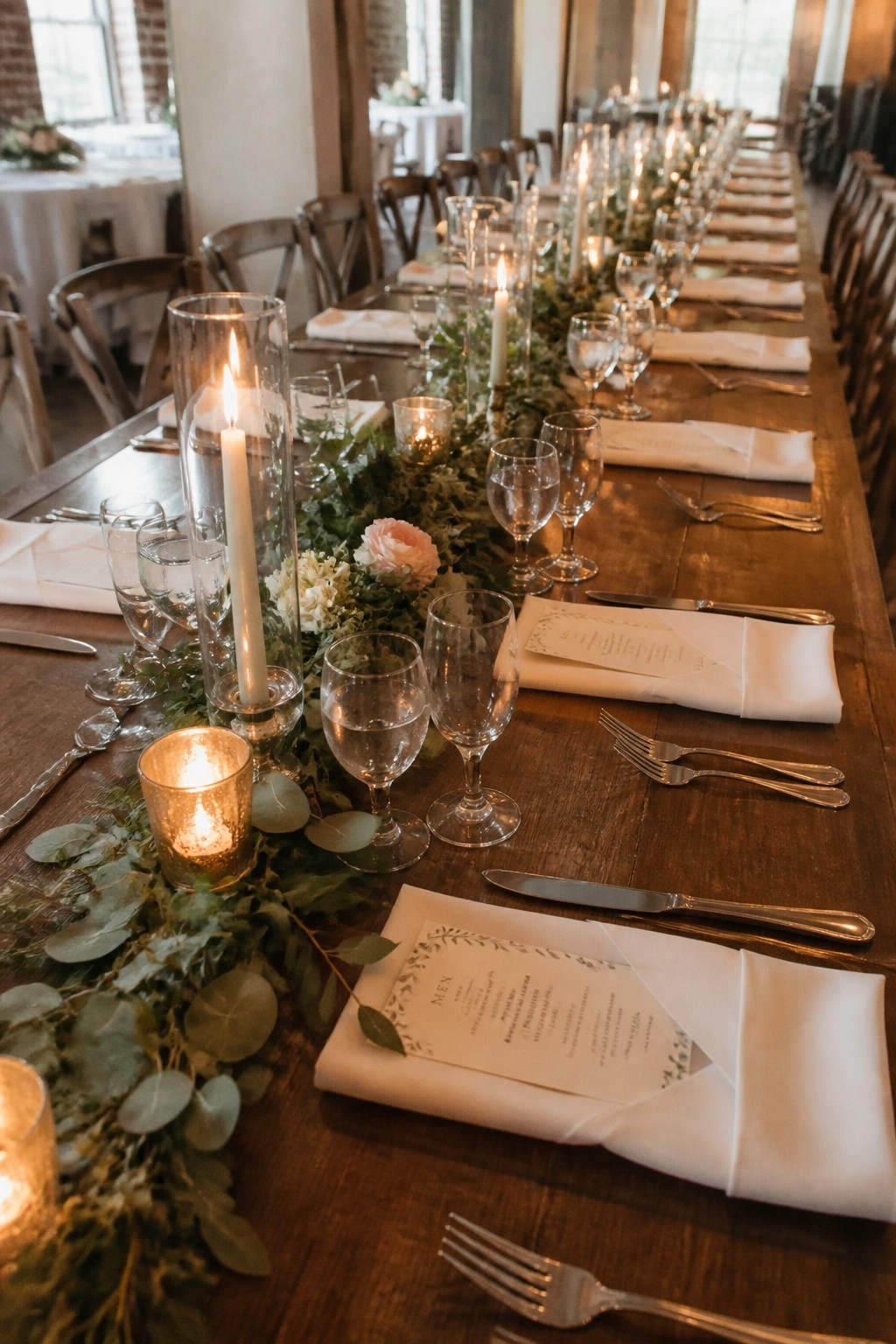 Long rustic wooden wedding table with eucalyptus and rose centerpiece, tall lit candles in glass holders, white folded napkins over menus, and formal glassware and silverware place settings.