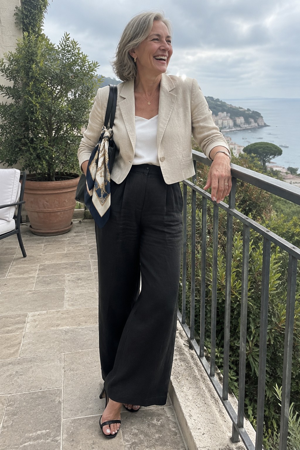 Gray-haired woman in beige blazer and black wide-leg pants smiles on seaside balcony.