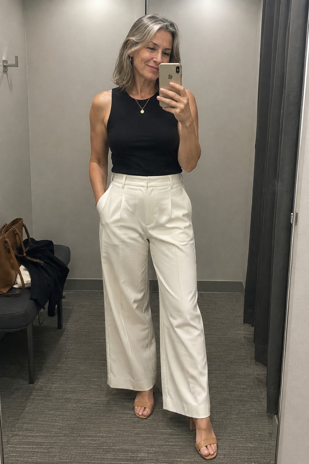 Woman with gray hair in black top and white pants taking mirror selfie
