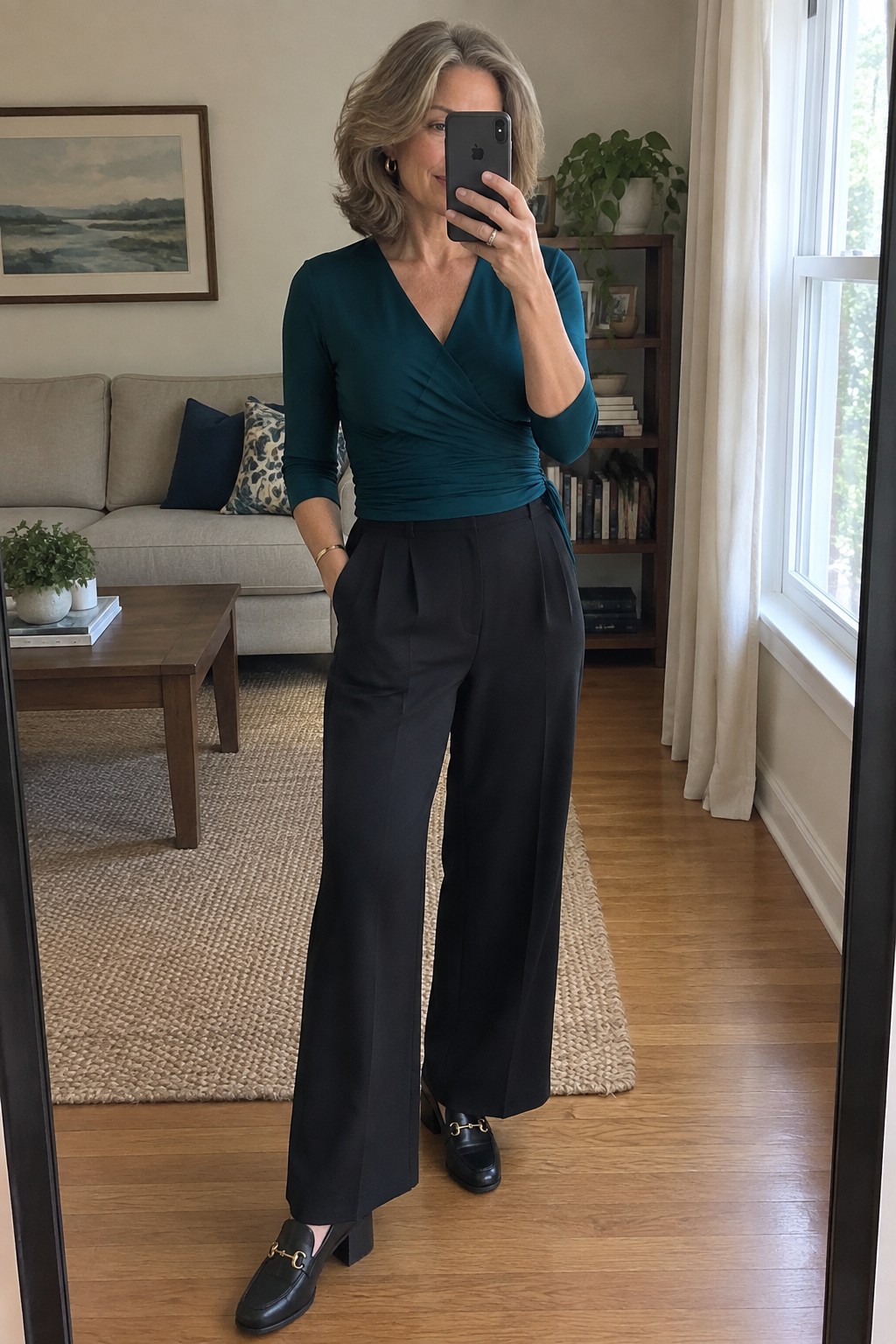 Gray-haired woman in teal wrap top and black wide pants taking mirror selfie