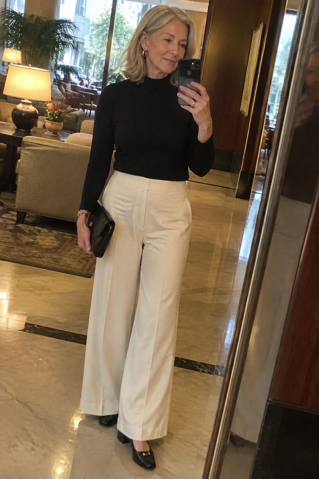 Woman in black turtleneck and white wide-leg pants taking mirror selfie in lobby