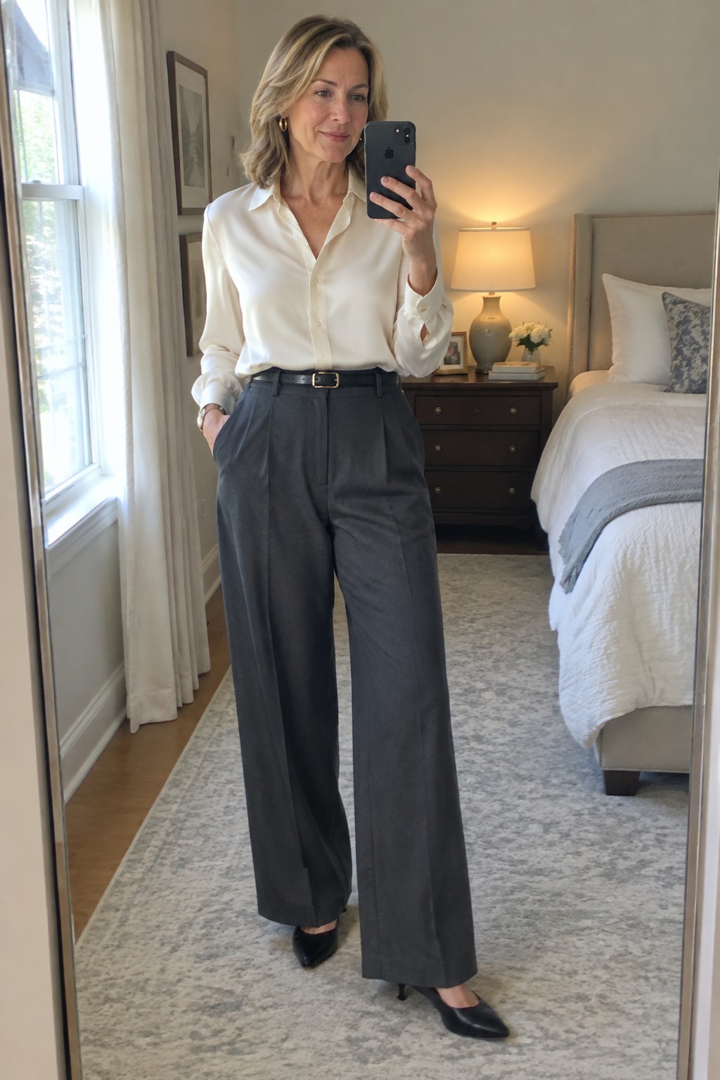 Woman in white shirt and gray trousers taking mirror selfie in bedroom