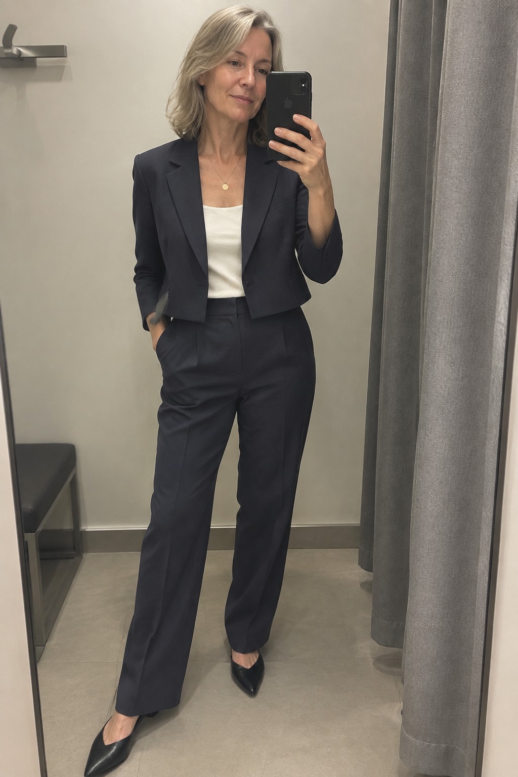 Woman in navy cropped suit holding iPhone for mirror selfie in fitting room