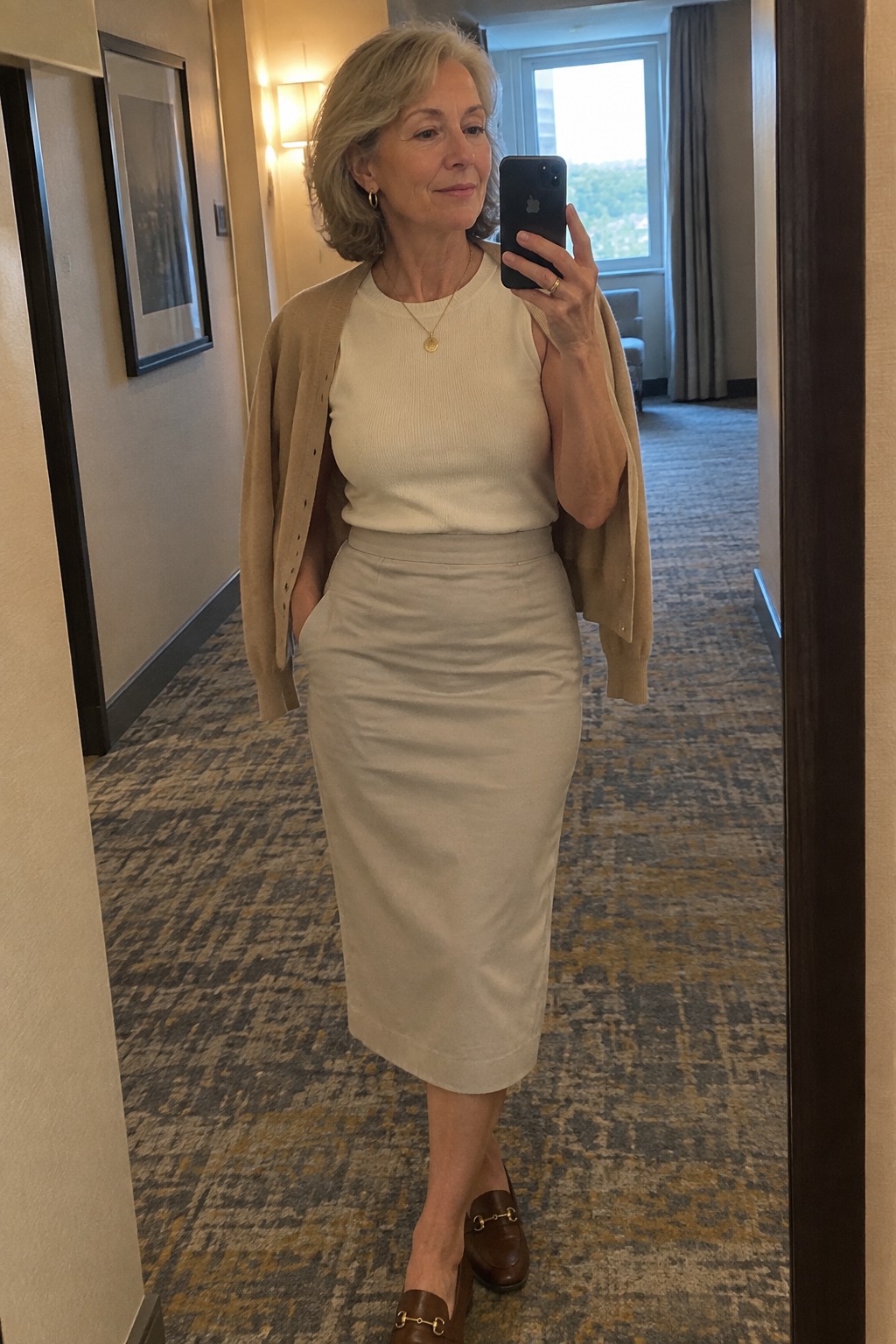 Silver-haired woman in cream top and skirt taking mirror selfie in hallway