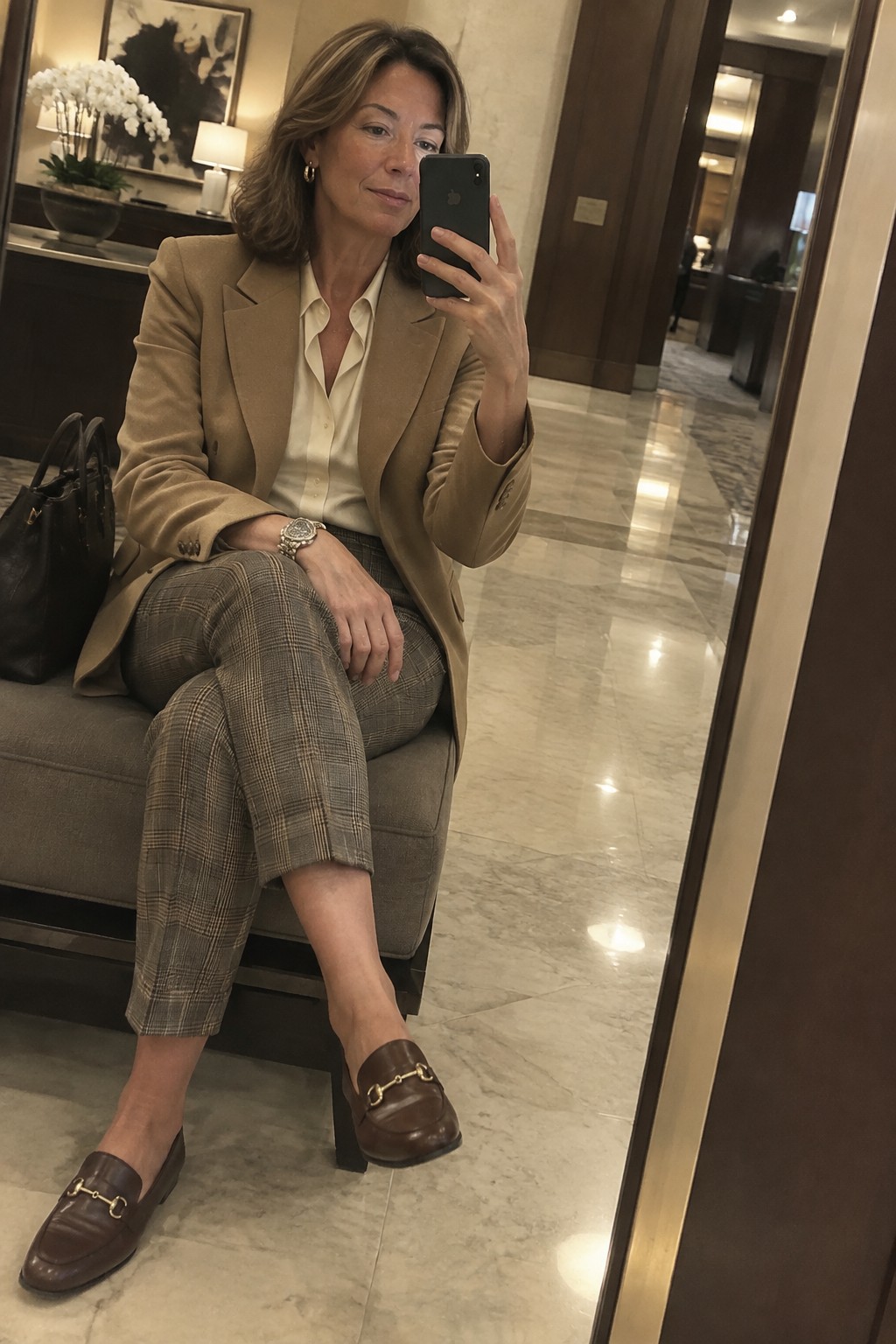 Woman in tan blazer over cream blouse with plaid cropped trousers and brown loafers holding a phone for a mirror selfie in a hotel lobby