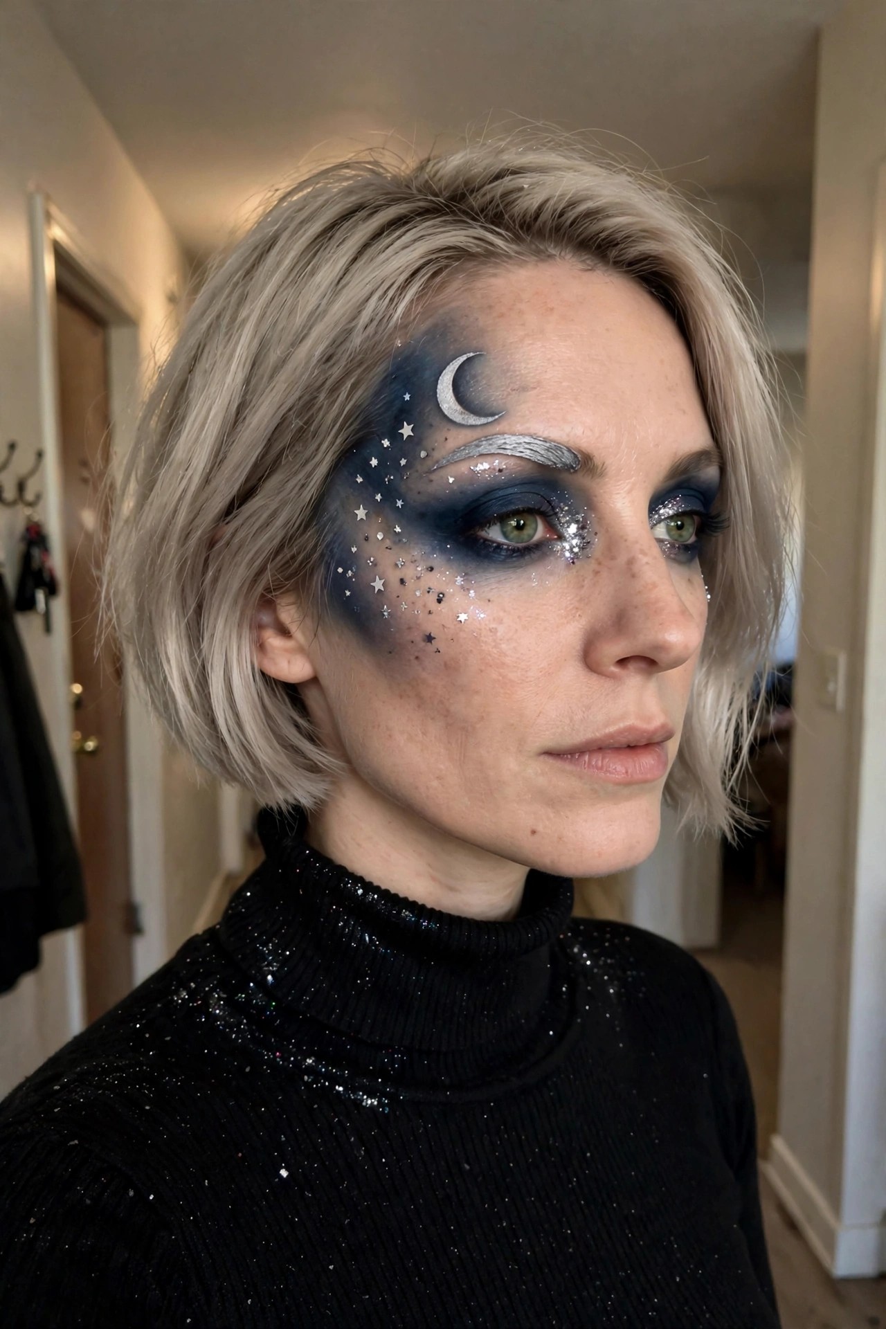Close-up of a woman with short blonde hair wearing dark blue smoky eye makeup, a silver crescent moon on the forehead, scattered silver stars across the temple and cheek, and glitter accents on the eye area.