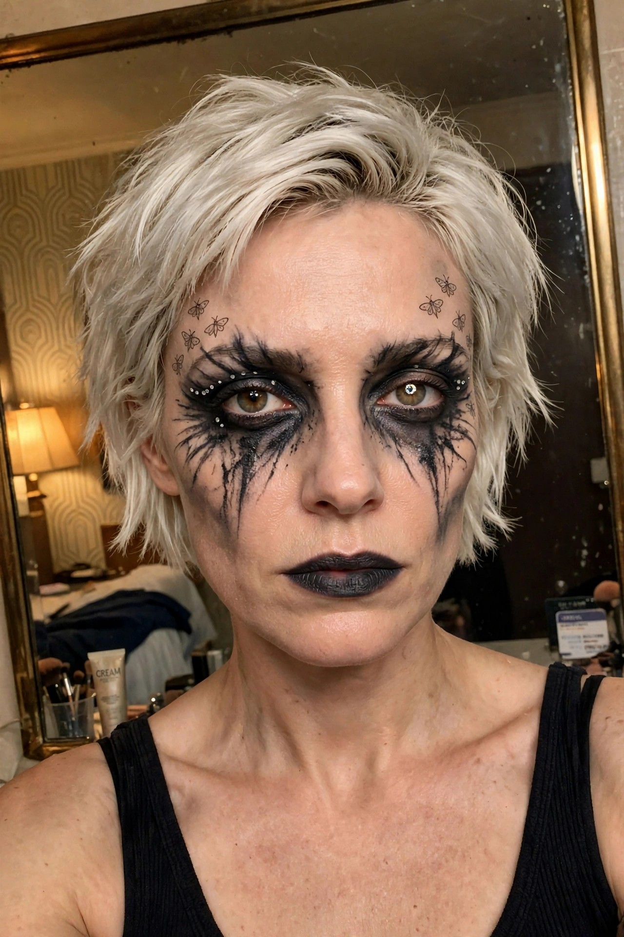 Close up of a person with short platinum hair wearing heavy black dripping eye makeup, small butterfly stamps on the forehead and temples, silver rhinestones around the eyes, and solid black lips.