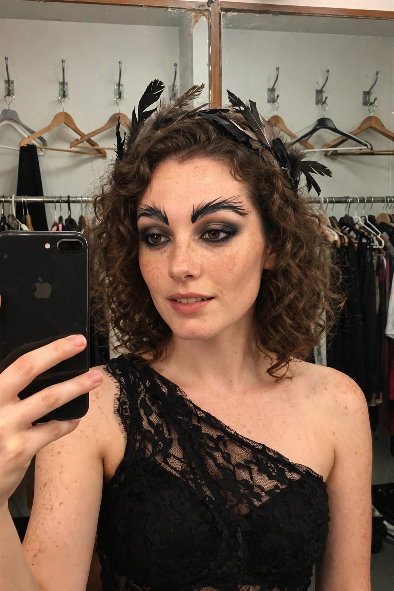 Woman wearing a black lace one-shoulder top and a black feather headpiece with dramatic black eye makeup and curly brown hair.