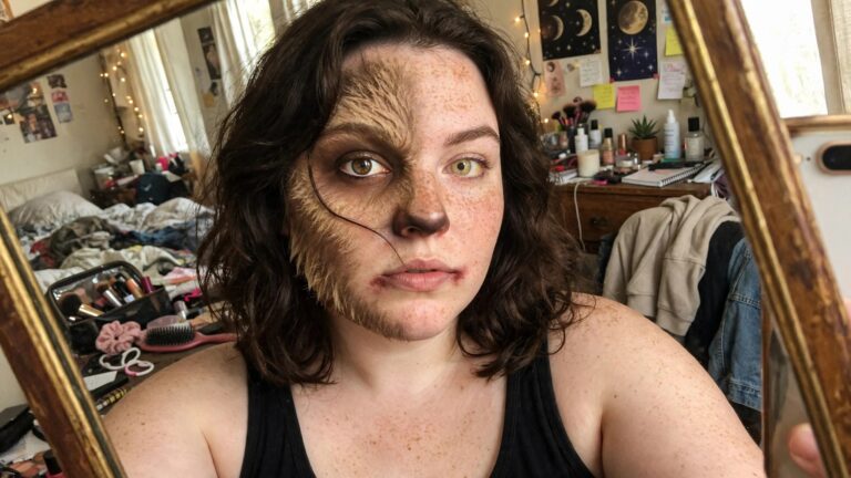 Close up of half face werewolf makeup with yellow contact lens, dark snout, and textured fur on the left side blending into natural freckled skin.