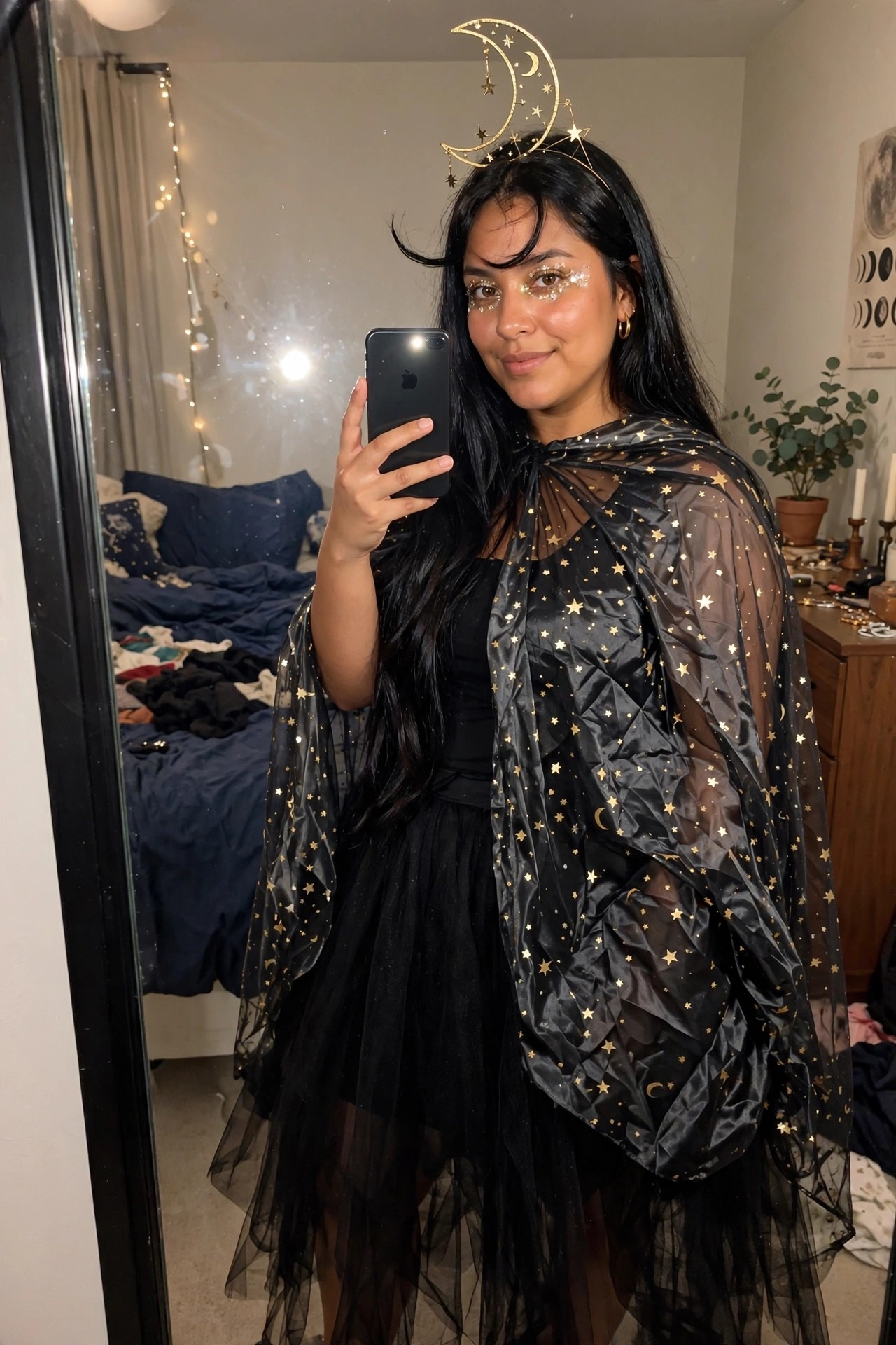 Woman wearing a sheer black cape printed with gold stars and moons paired with a crescent moon headpiece and glitter eye makeup for a celestial witch costume.