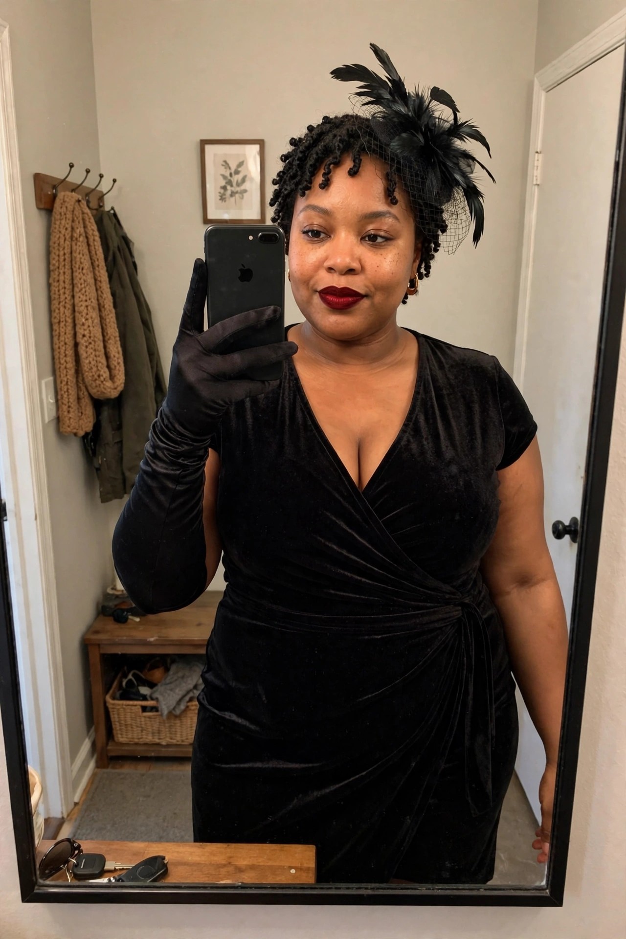 Woman wearing a black velvet wrap dress with long black gloves and a black feathered fascinator with veil netting, taking a mirror selfie.