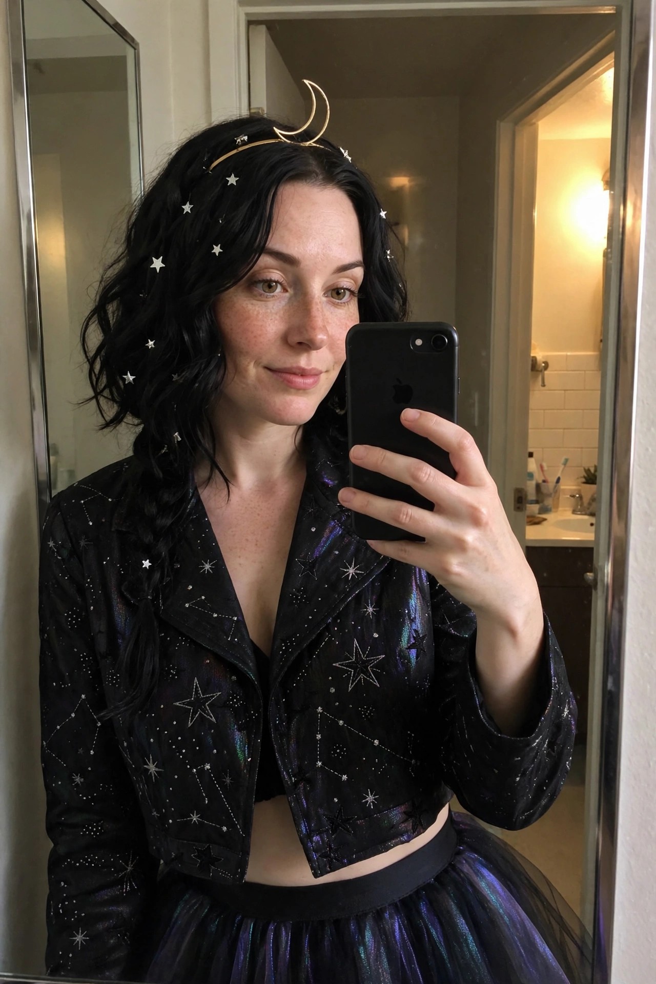 Woman wearing a black cropped jacket with constellation prints and stars, paired with an iridescent black tulle skirt, gold crescent moon headband, and small star clips in long black hair.