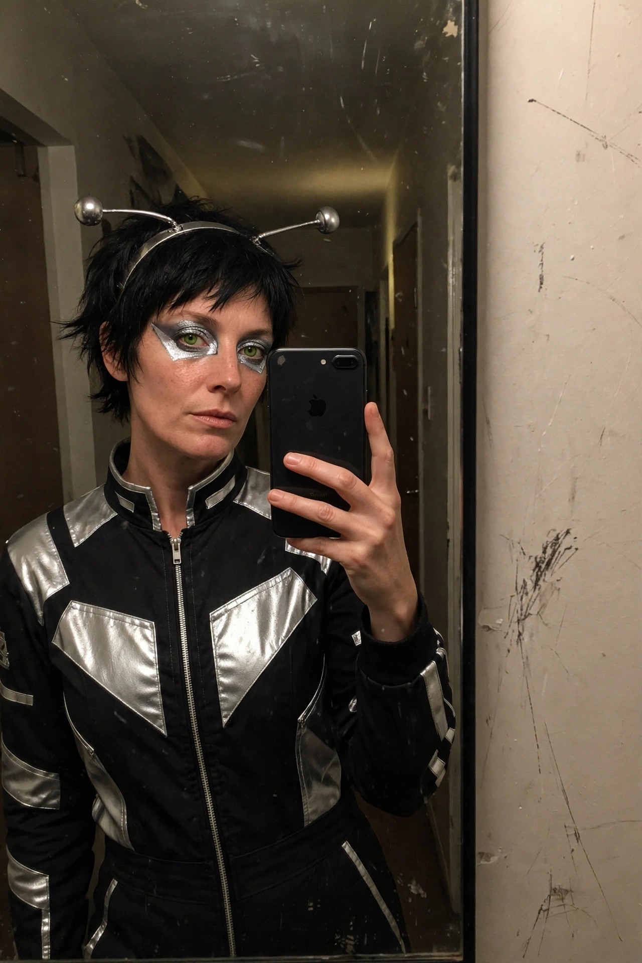 Person wearing a black and silver metallic jumpsuit with a silver antenna headpiece and silver eye makeup, short black hair visible.
