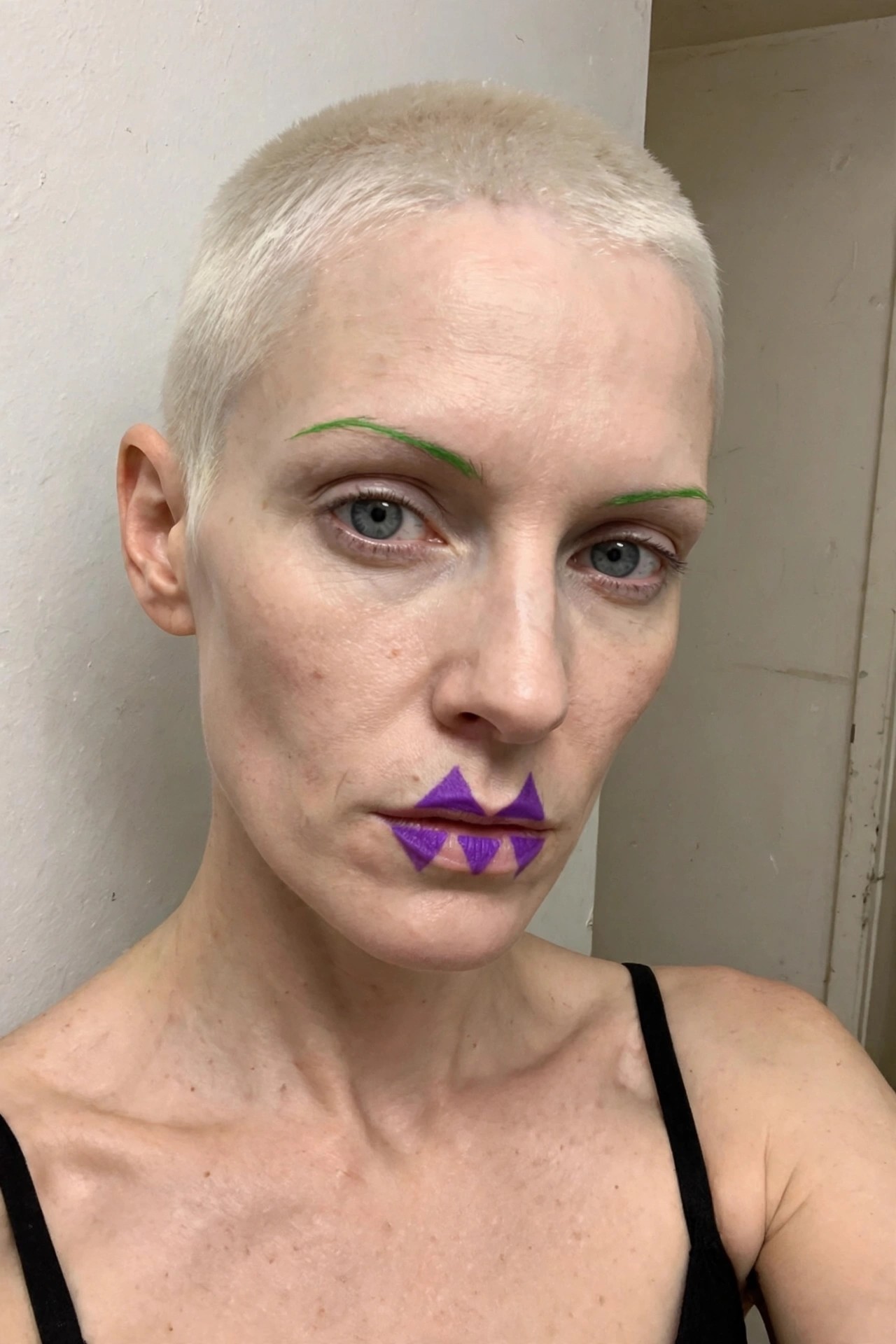 Close-up of Joker-inspired makeup with purple triangular shapes painted around the lips and bright green drawn-on eyebrows on a person with short bleached hair.