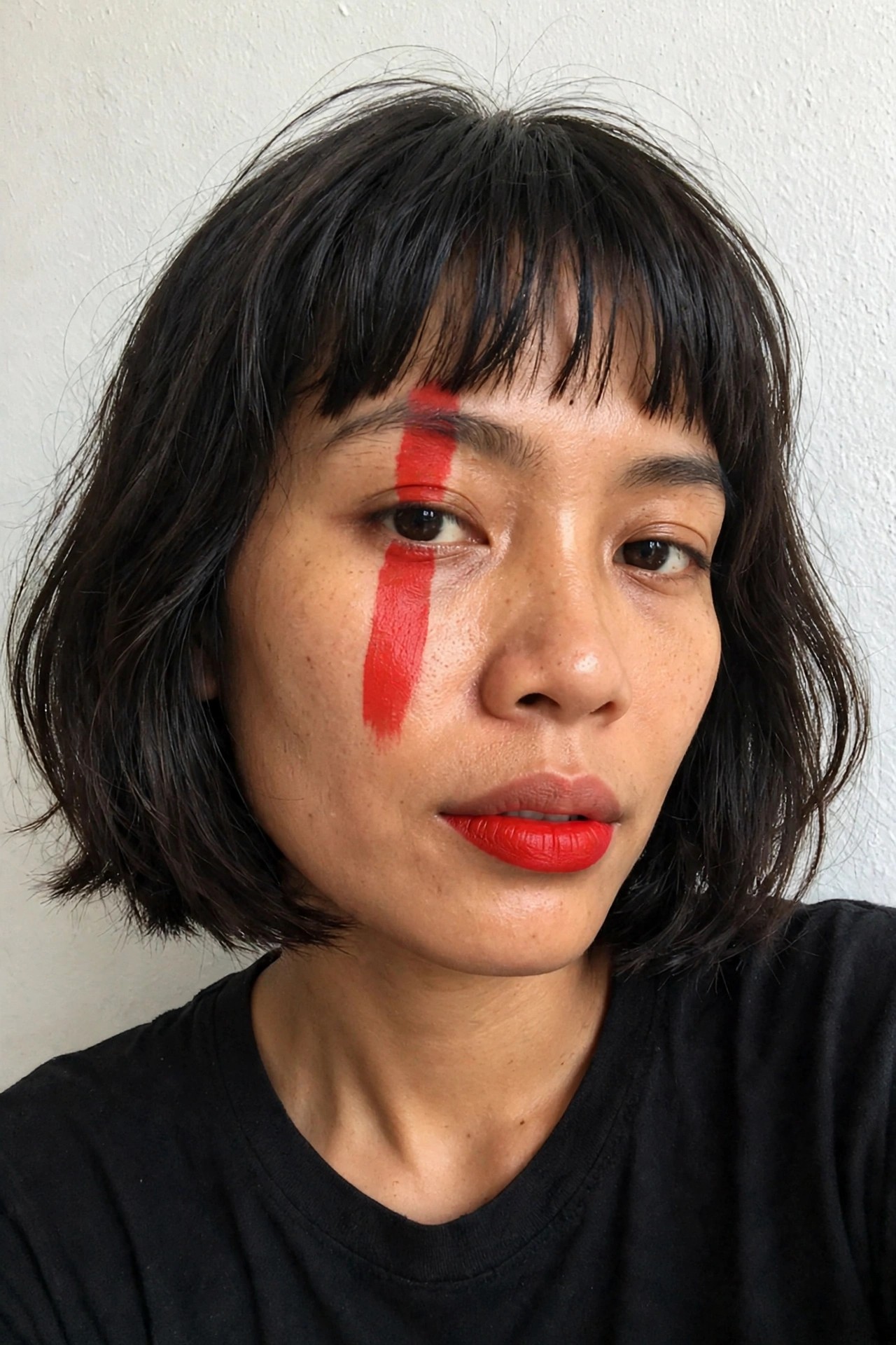 Close up of a woman with short dark hair wearing red lipstick and a thick vertical red stripe painted down the center of her face from forehead to cheek.