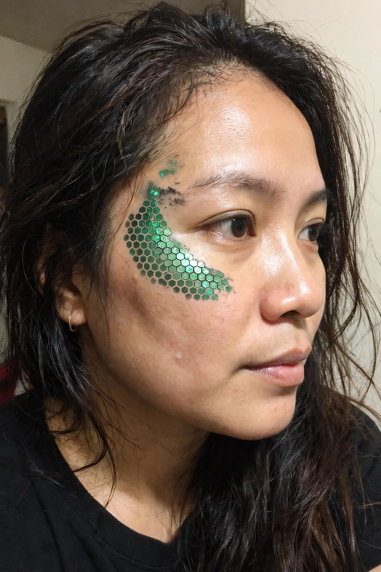 Close up of a woman wearing green hexagonal sequins arranged in overlapping scale rows around one eye with scattered loose glitter above the brow.