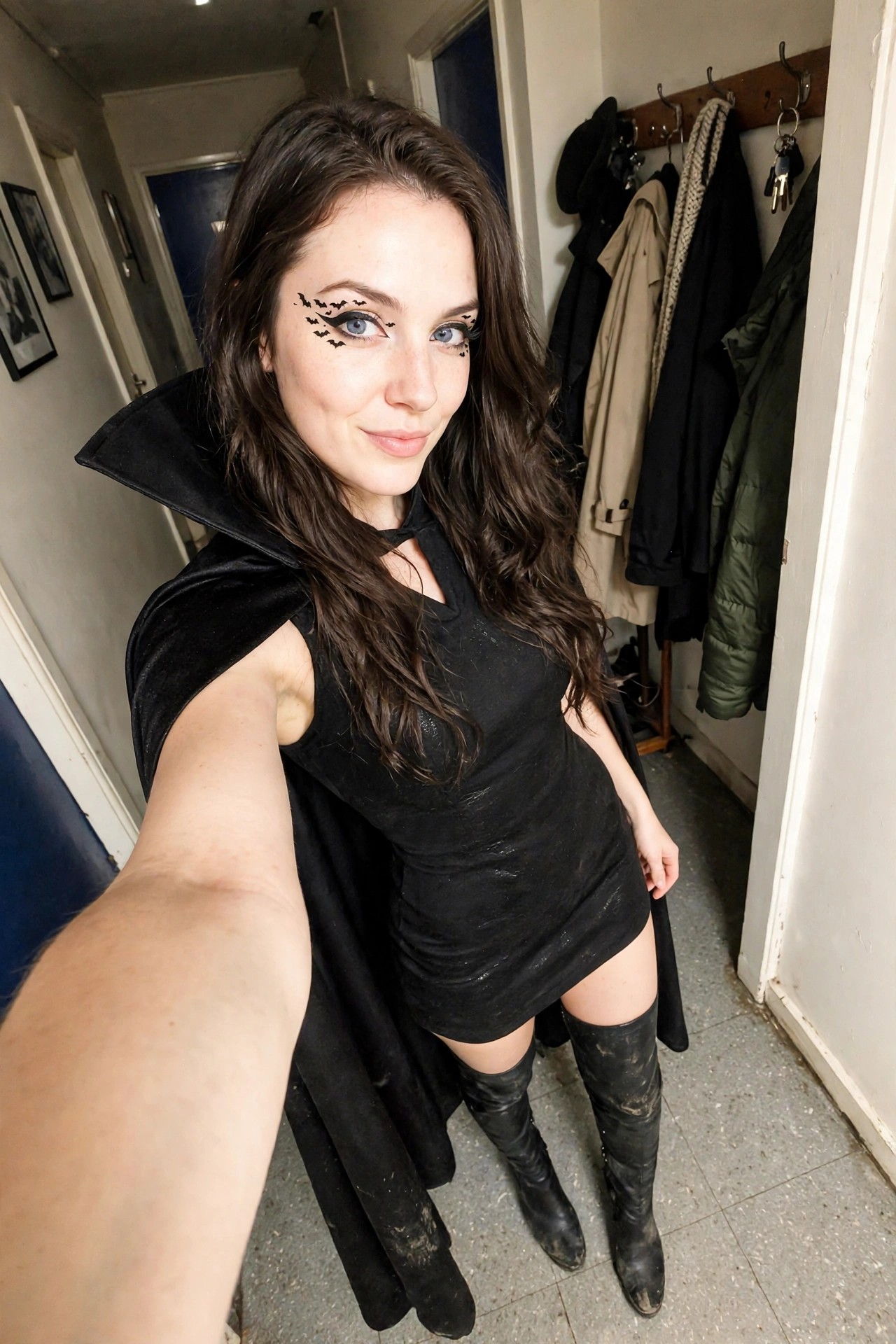 Woman wearing a black mini dress with a large black velvet cape, black boots, and bat decals around her eyes as vampire makeup.